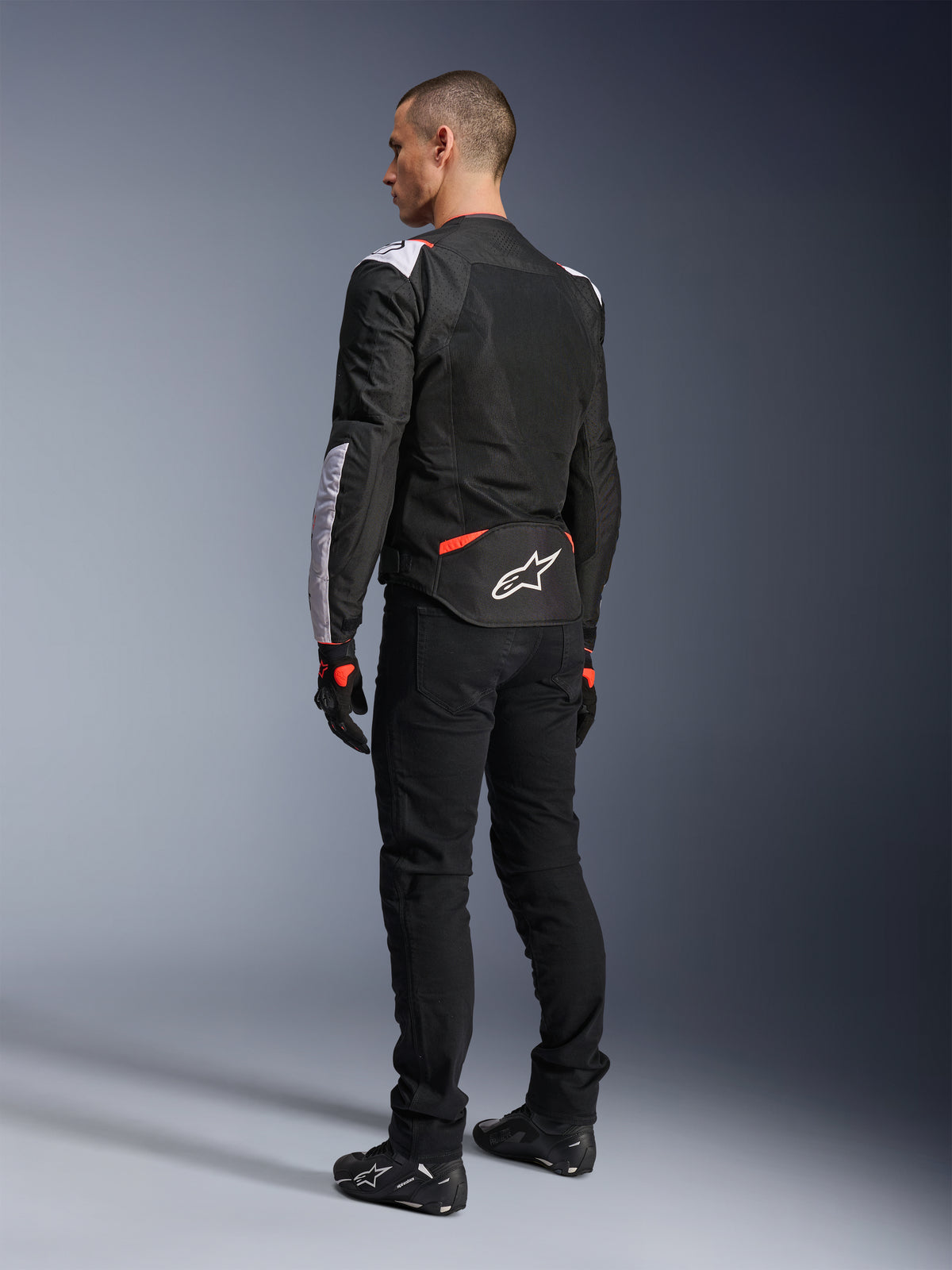 Giacca Alpinestars T-Sps Air V2, giacca da moto in tessuto, nero, bianco e rosso fluo, vista posteriore su modello, pannelli in mesh per la ventilazione, logo Astars bianco sulla parte inferiore della schiena, abbinata a pantaloni e guanti da moto neri