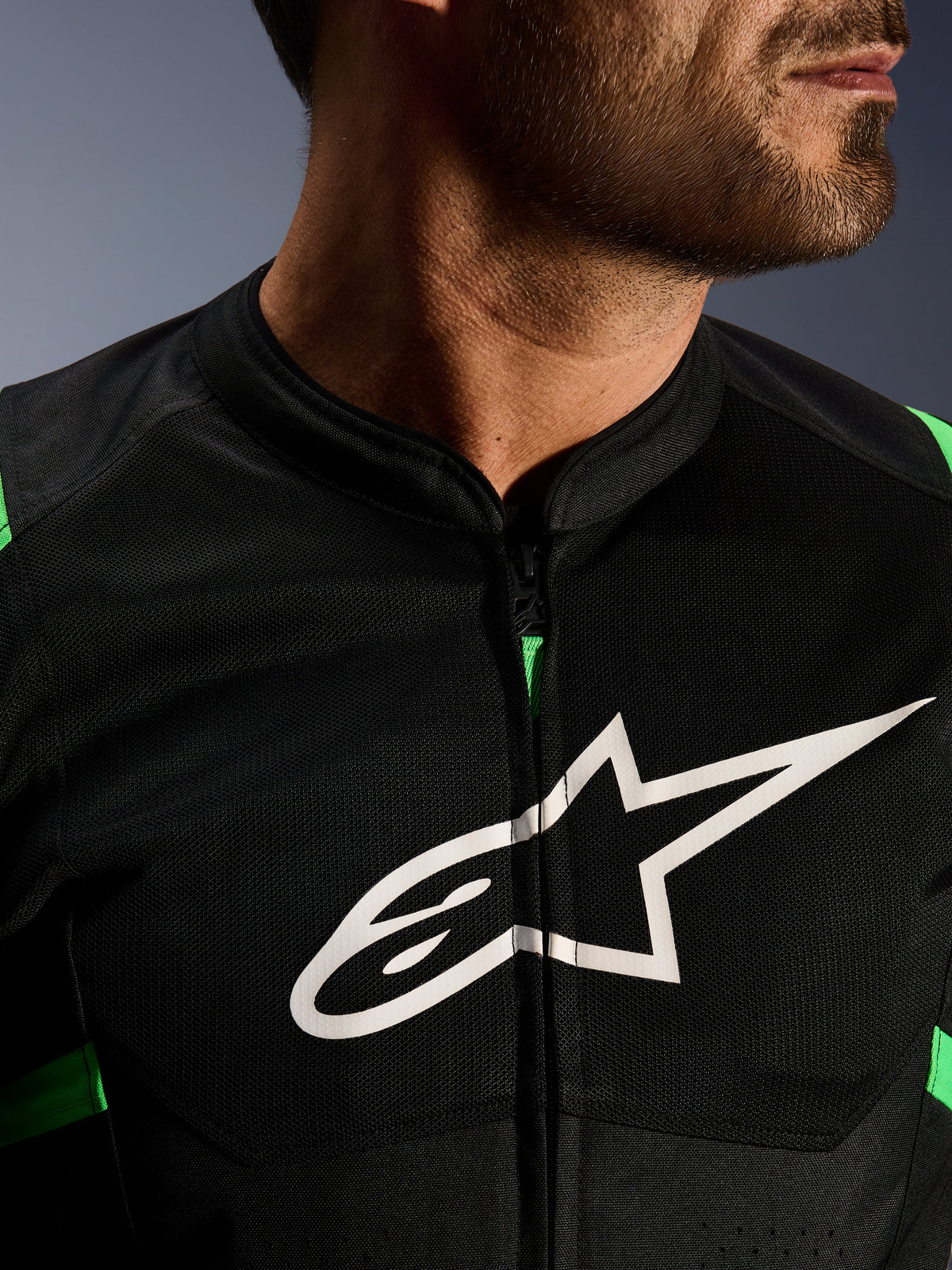 Primo piano della giacca Alpinestars T-SPS Air V2, nero e verde fluo, con pannelli in rete tecnica traspirante, cerniera centrale e prominente logo a stella Alpinestars bianco sul petto, abbigliamento tecnico da moto