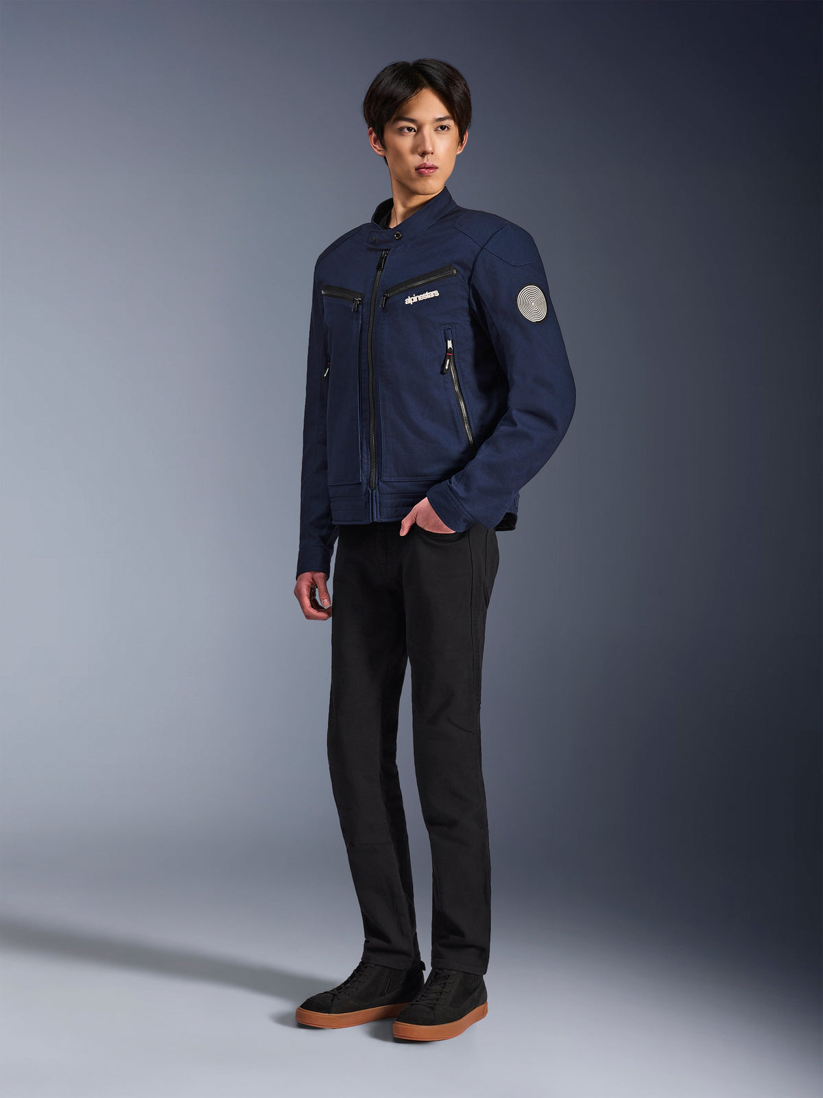 Giacca Alpinestars Bruiser in blu scuro, giacca da moto urbana in tessuto dal design d'ispirazione workwear, con tasche sul petto con cerniera e patch Oscar a spirale sulla manica, indossata con pantaloni neri e sneakers casual.