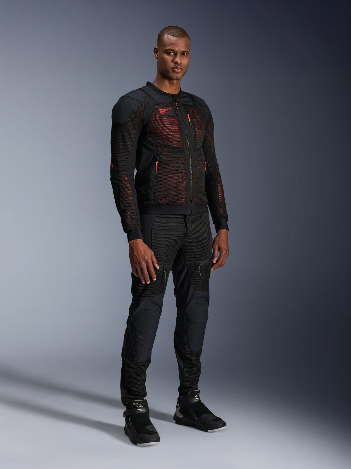 Giacca Alpinestars Troop-Air Pro in Nero Rosso Fluo, giacca da moto tecnica in mesh corazzata indossata da un modello uomo con pantaloni neri corazzati coordinati e stivali alti, ritratto in studio a figura intera.