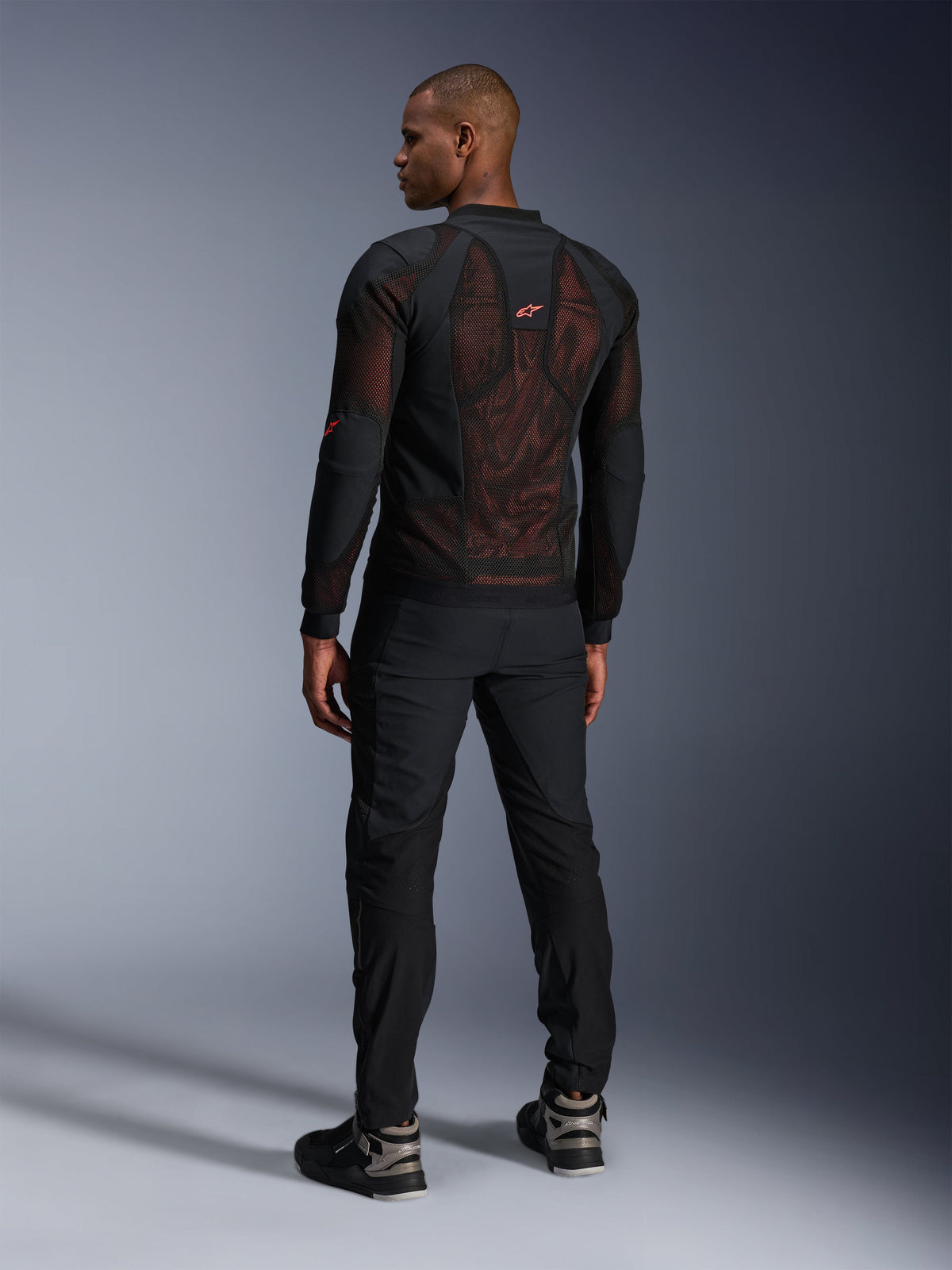 Giacca Alpinestars Troop-Air Pro in Black Red Fluo, vista posteriore su un modello, con un ampio pannello posteriore in mesh tecnica 2D e interno rosso visibile, abbinata a pantaloni tecnici neri e stivali neri e grigi, design moderno di giacca da moto urbana protettiva.