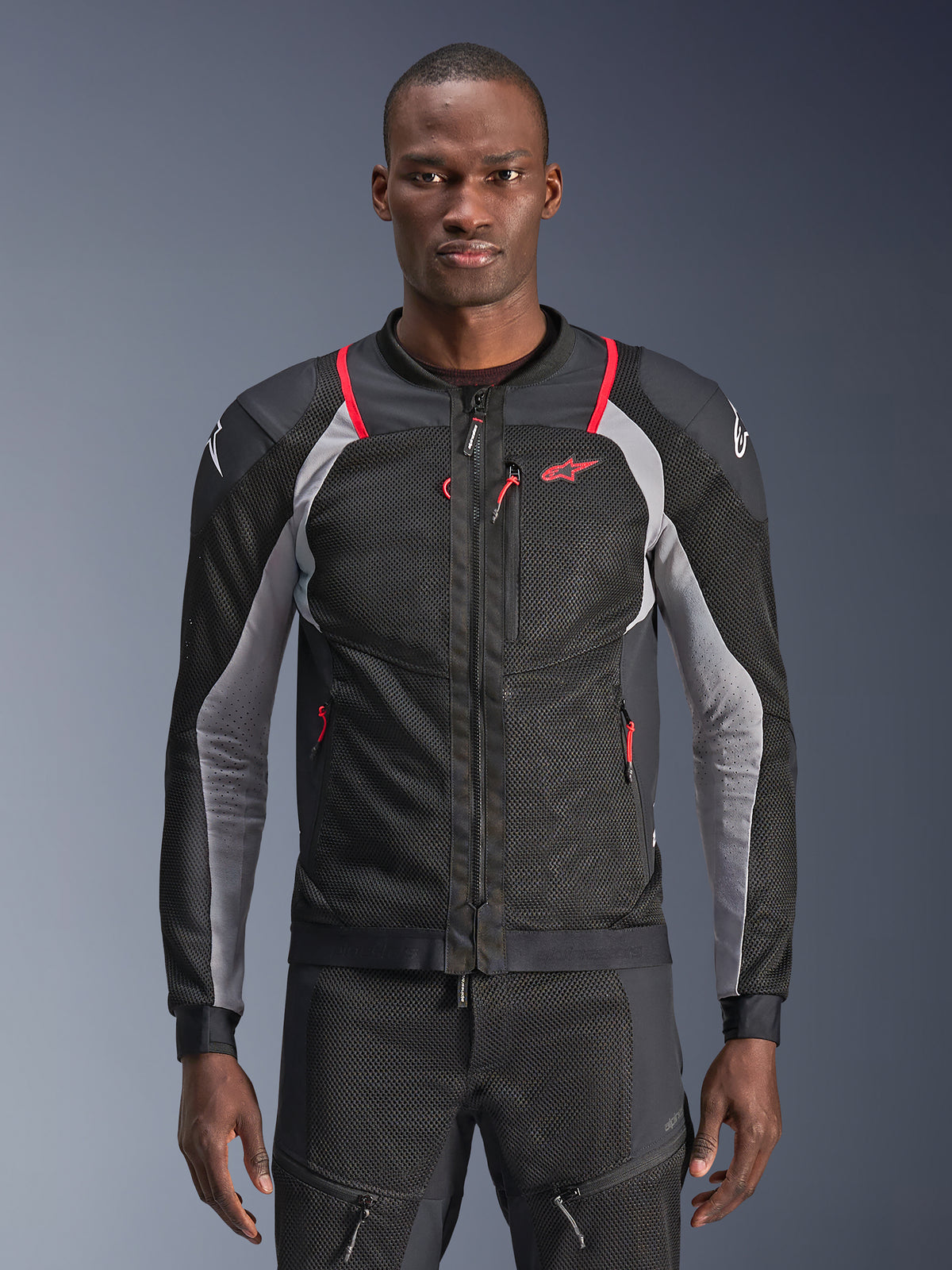 Alpinestars Troop-Air Pro Jacket, nero, grigio cenere e rosso brillante, indossata da un modello, giacca da moto in mesh con pannelli di ventilazione traforati, con tasche con cerniera rosse e loghi bianchi sulle spalle, abbigliamento tecnico protettivo