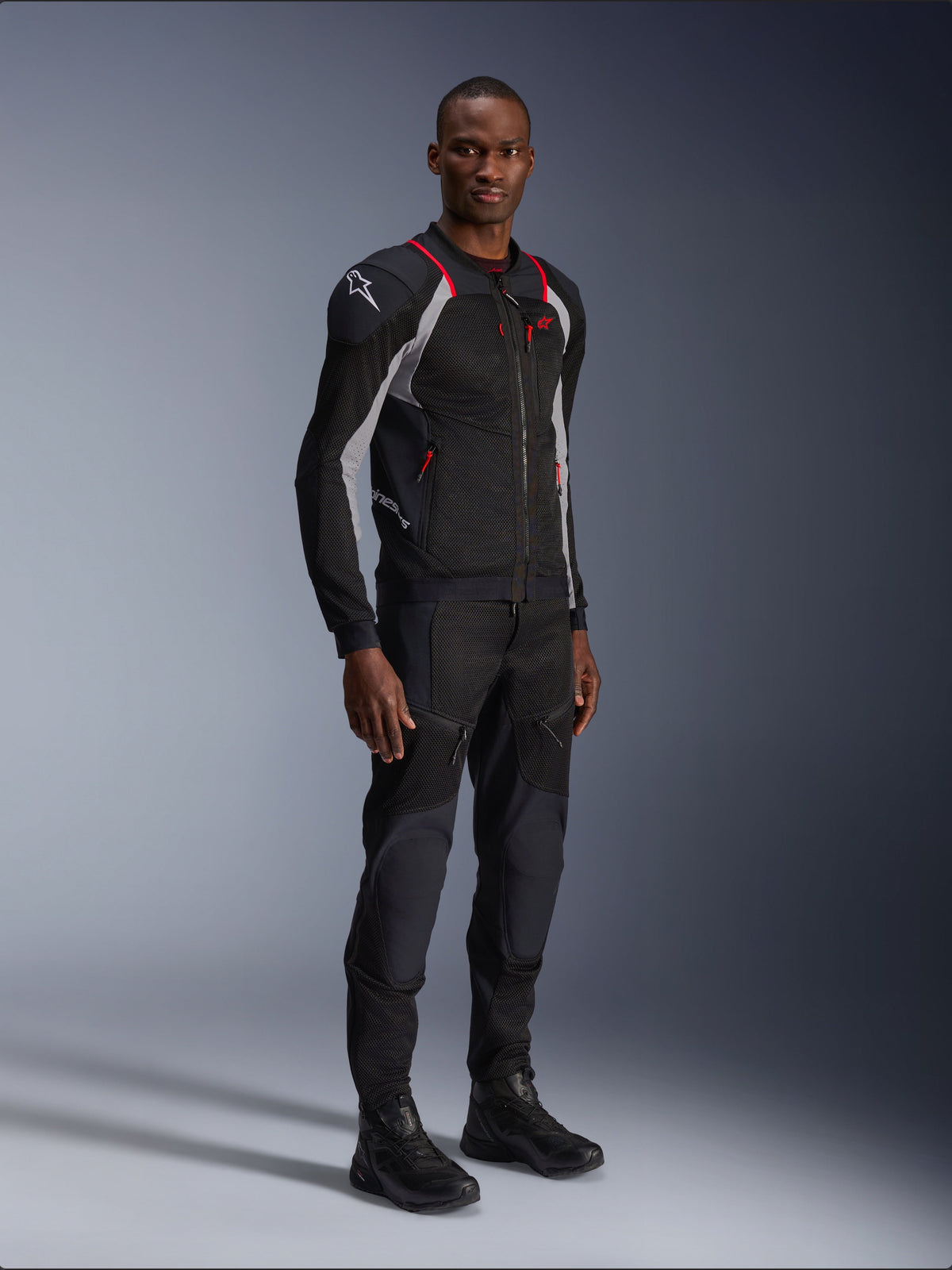 Giacca Alpinestars Troop-Air Pro, nero grigio cenere e rosso brillante, giacca tecnica a rete corazzata, con accenti rossi e logo bianco sulla spalla, indossata con pantaloni e stivali neri coordinati, abbigliamento da moto urbano moderno