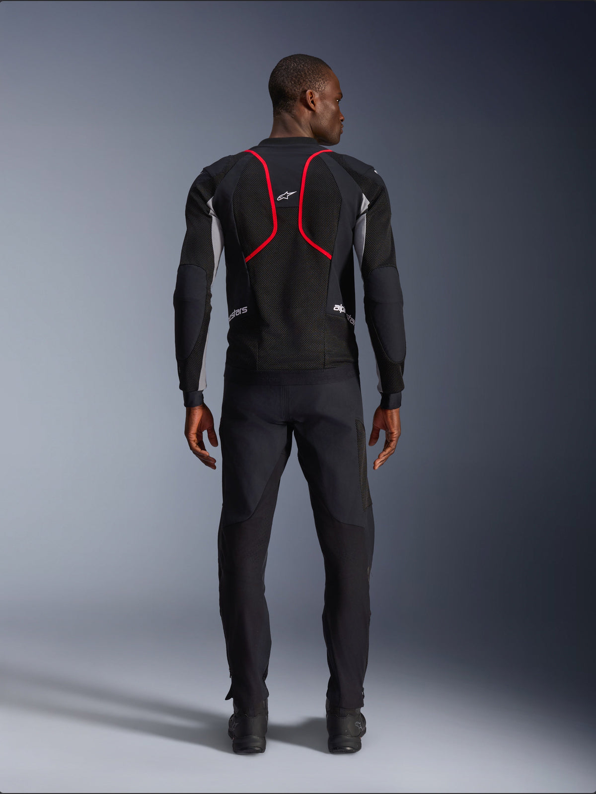Modello visto da dietro che indossa la giacca Alpinestars Troop-Air Pro, nero con accenti grigio cenere e rosso acceso, caratterizzata da una costruzione in mesh tecnico con evidenti profili rossi sulla schiena, logo Alpinestars sulla parte superiore della schiena, abbinata a pantaloni da moto neri, giacca tecnica ventilata premium per guida su strada