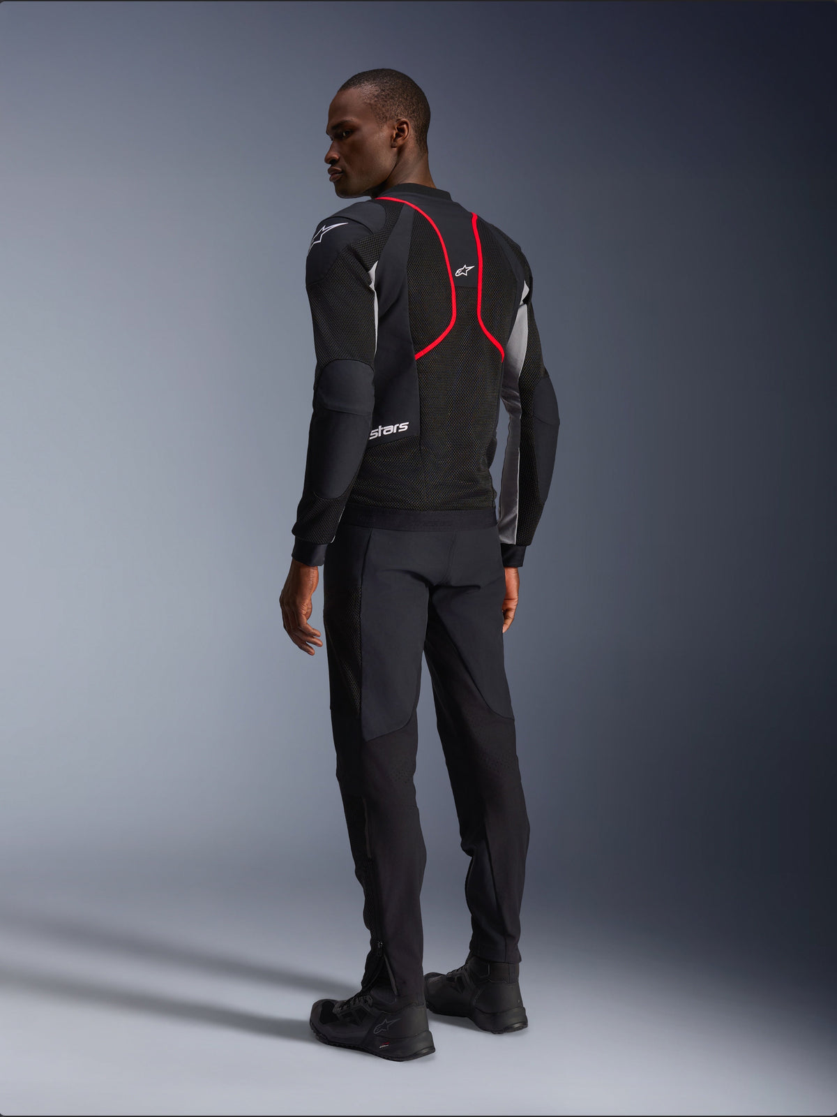 Alpinestars Troop-Air Pro Jacket, giacca da moto, nero, grigio cenere e rosso vivace, vista posteriore su modello, con linee d'accento rosse e pannelli di ventilazione in mesh, indossata con pantaloni e stivali neri, stile motociclistico urbano
