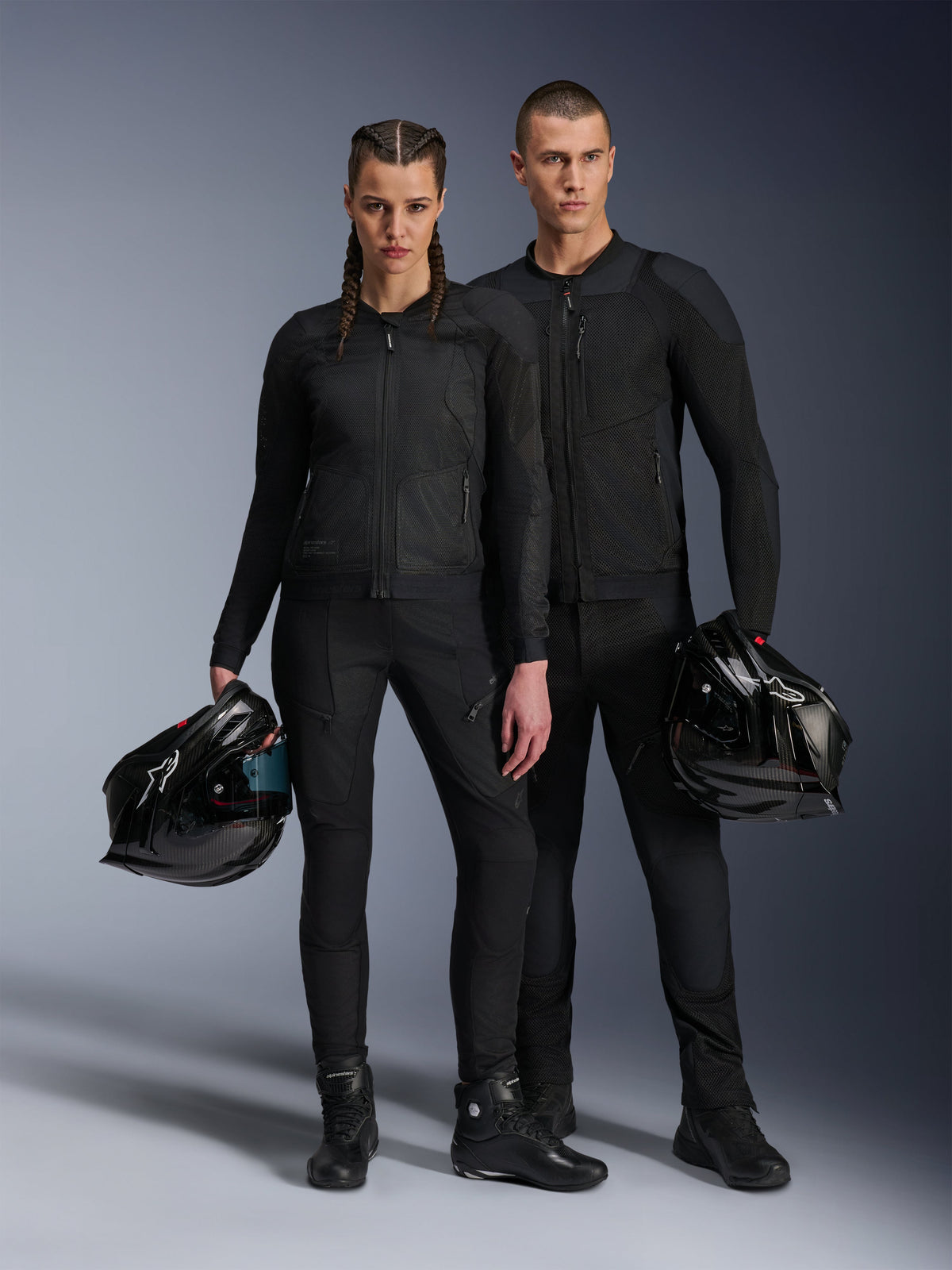 Modelli uomo e donna che indossano giacche Alpinestars Troop-Air Pro nere, abbigliamento tecnico da moto corazzato in mesh abbinato a pantaloni e stivali neri coordinati, ciascuno con un casco nero, per un look da moto urbano coordinato.
