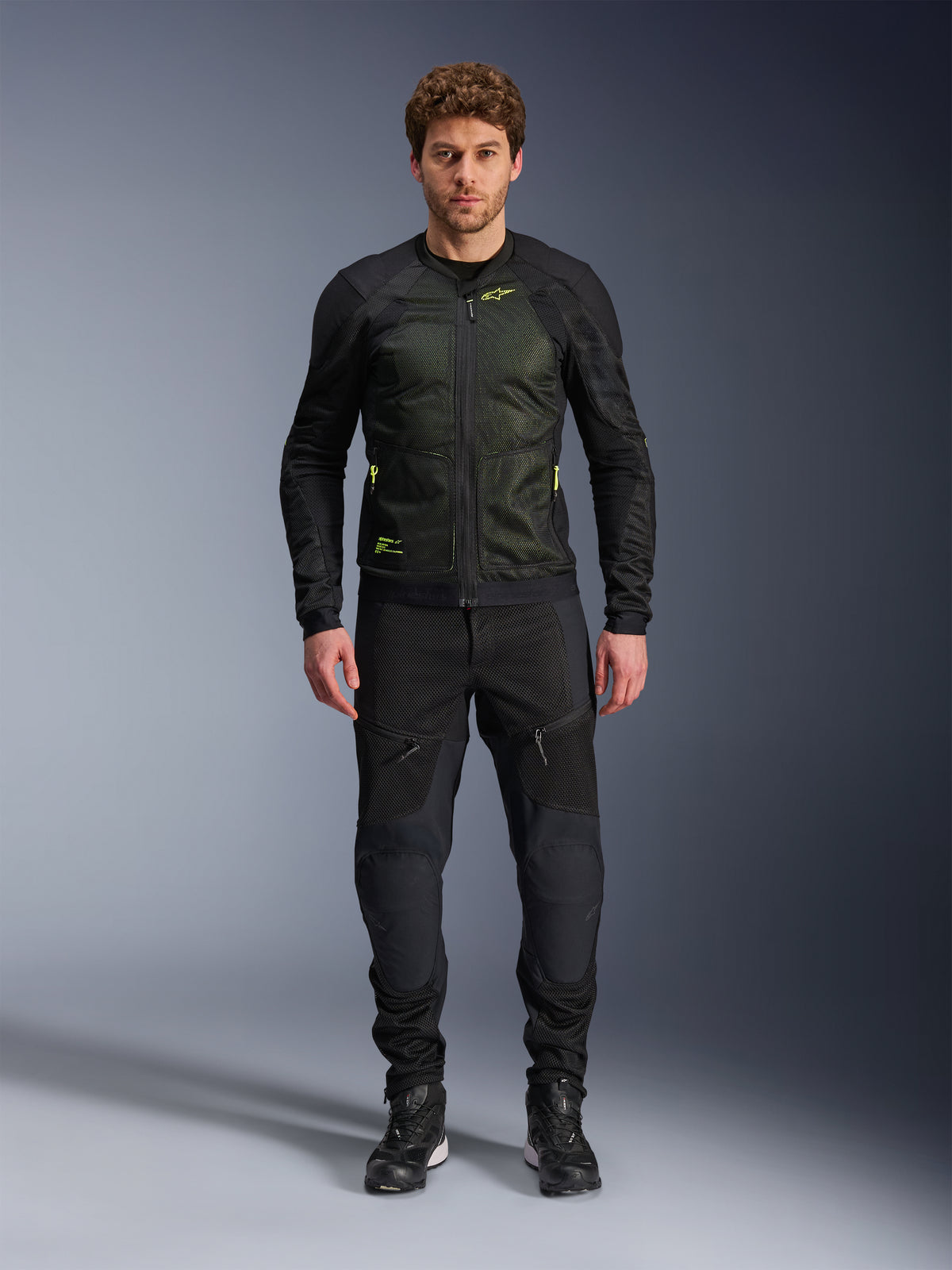 Modello che indossa la giacca Alpinestars Troop-Air in Nero Giallo Fluo, con pannelli in mesh tecnico e accenti giallo brillante, abbinata a pantaloni da moto e scarpe neri, abbigliamento da moto contemporaneo per climi caldi