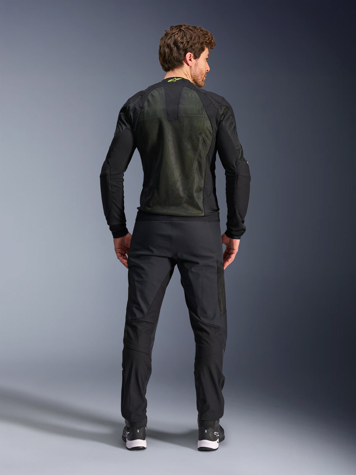 Giacca Alpinestars Troop-Air, Nero Giallo Fluo, vista posteriore su un modello, con ampio pannello posteriore in mesh tecnico 2D per la ventilazione, giacca tecnica da moto indossata con pantaloni da moto e stivali neri, stile motociclistico urbano