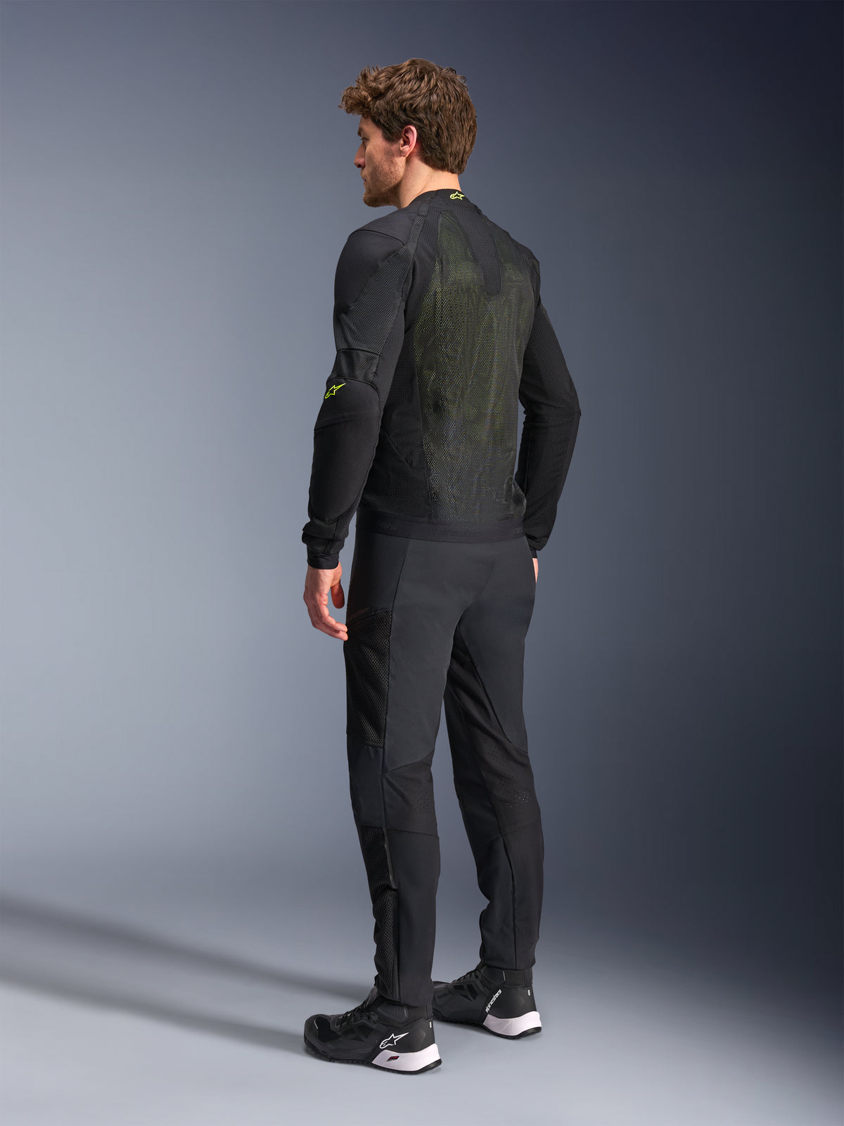 Giacca Alpinestars Troop-Air in nero e giallo fluo, vista posteriore di un modello che indossa la giacca tecnica, ampio pannello posteriore in mesh con riflessi gialli e loghi Alpinestars giallo fluo, abbinata a pantaloni da moto neri