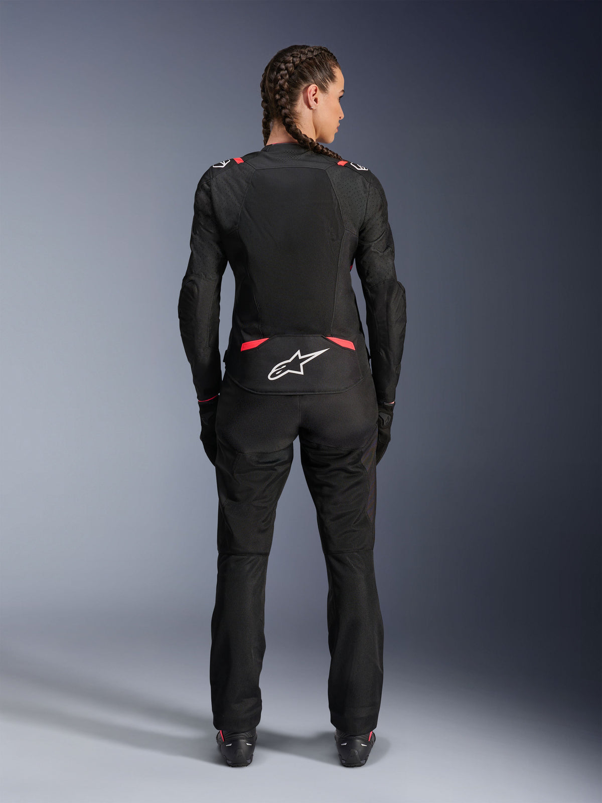 Alpinestars Stella T-SPS Air V2 Jacket, giacca da moto in tessuto da donna, nero e rosa Diva, vista posteriore su modella con capelli intrecciati, con ampi pannelli in mesh per la ventilazione, logo Astars bianco sulla parte inferiore della schiena, abbinata a pantaloni da moto neri coordinati.