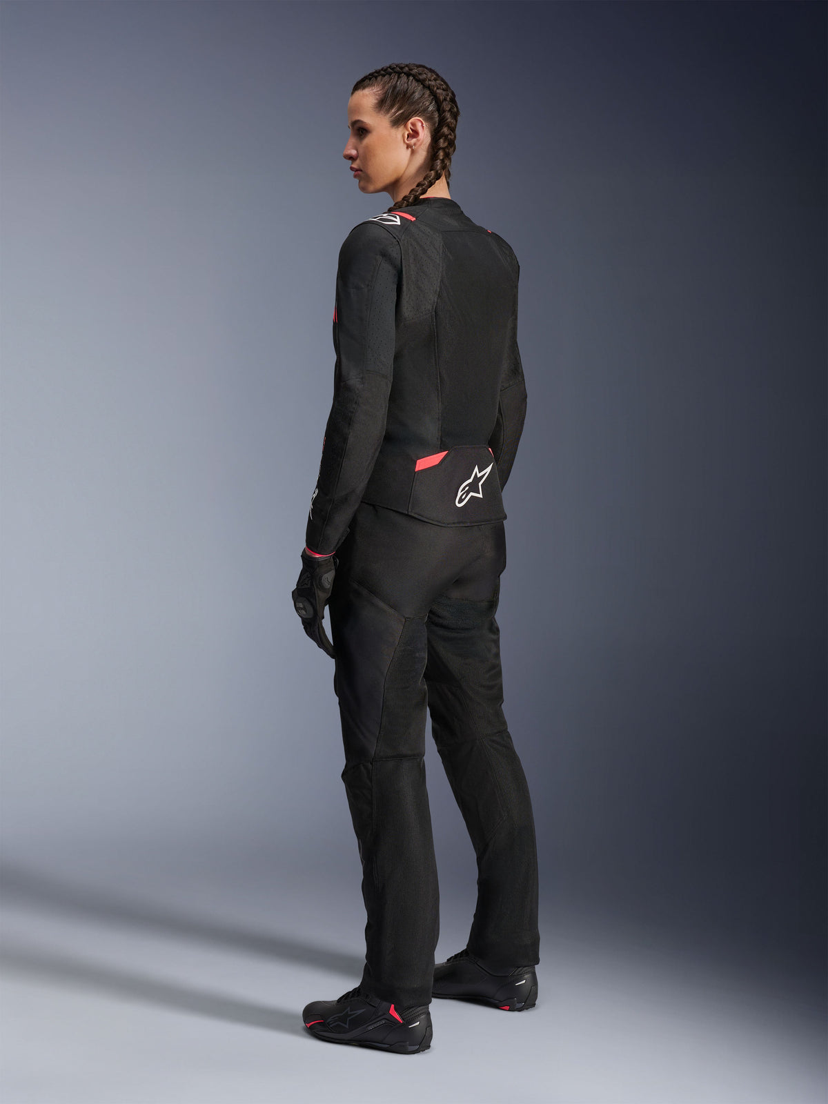 Giacca Alpinestars Stella T-Sps Air v2 da donna, nero e Diva Pink, giacca da moto in tessuto vista da una prospettiva posteriore-laterale, con ampi pannelli in mesh per il flusso d'aria e logo a stella bianco sulla schiena, indossata con pantaloni da moto, guanti e stivali coordinati neri.