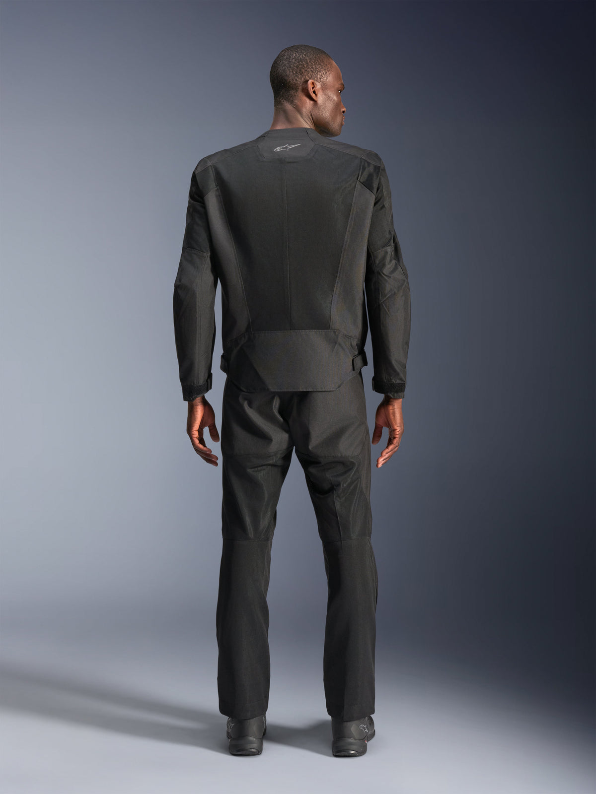 Vista posteriore di un uomo che indossa la giacca Alpinestars C-1 Air in nero, giacca da moto urbana con pannelli in rete tecnica, pantaloni in tessuto e stivali neri coordinati, estetica pulita con logo riflettente sulla parte superiore della schiena.