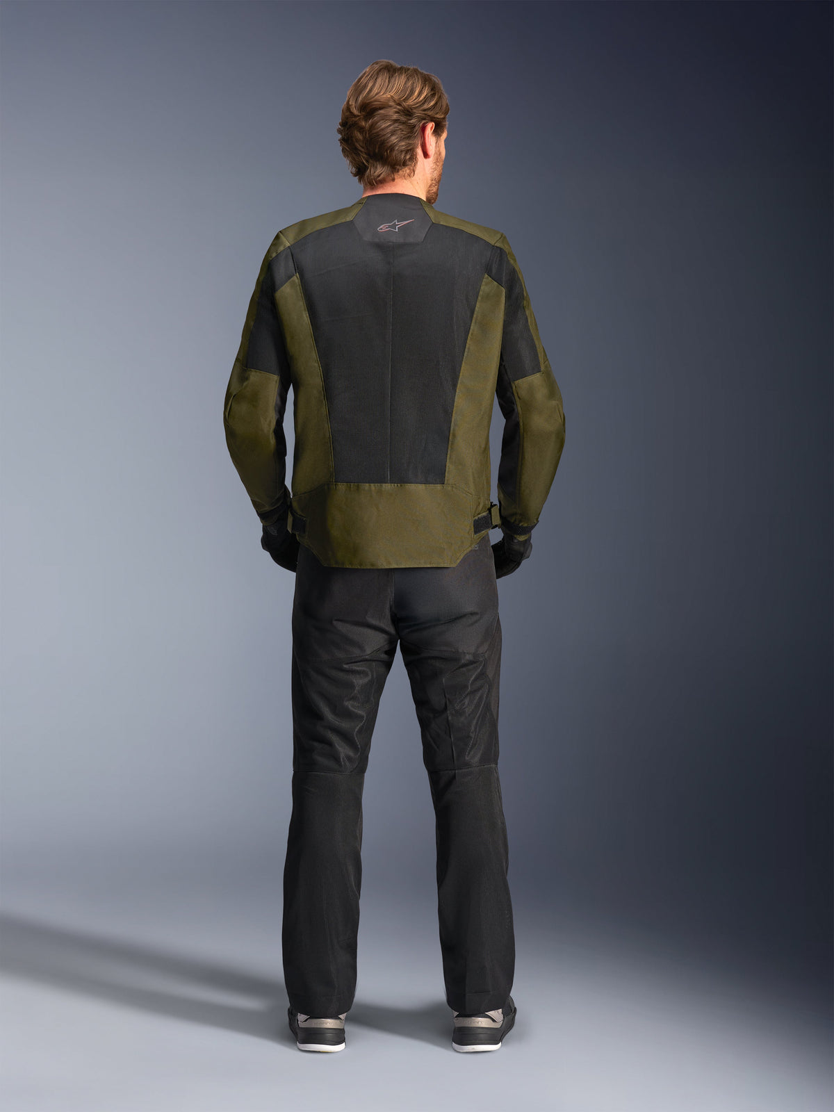 Vista posteriore di un motociclista che indossa la giacca Alpinestars C-1 Air in kaki e nero, con un ampio pannello posteriore in rete tecnica per la ventilazione, abbinata a pantaloni da moto neri, abbigliamento urban contemporary per guida su strada.