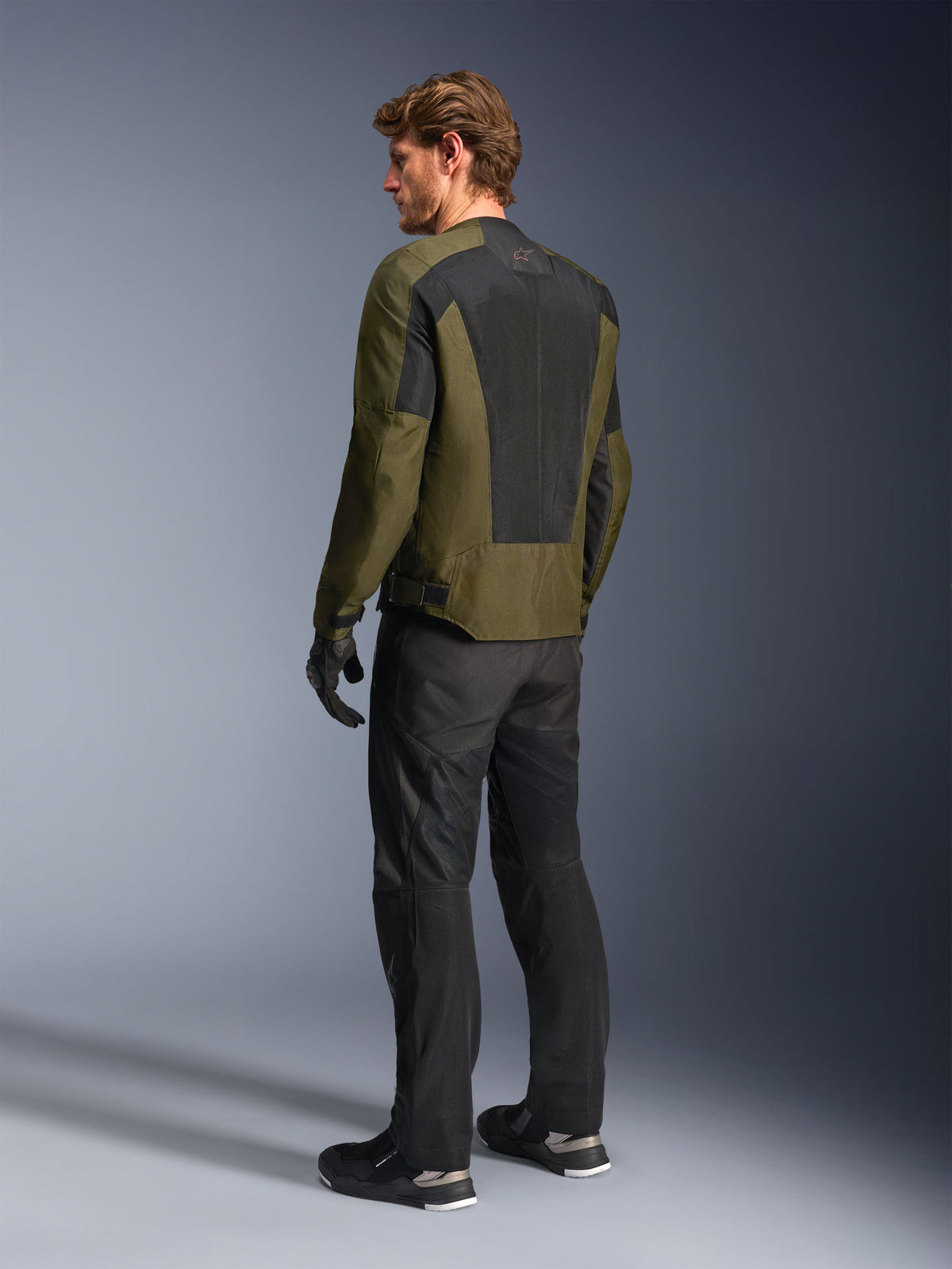 Modello uomo con giacca Alpinestars C-1 Air in colore Khaki, giacca da moto urban, vista posteriore con ampi pannelli in rete nera sulla schiena, abbinata a pantaloni tecnici da guida e guanti neri, abbigliamento da moto contemporaneo per climi caldi