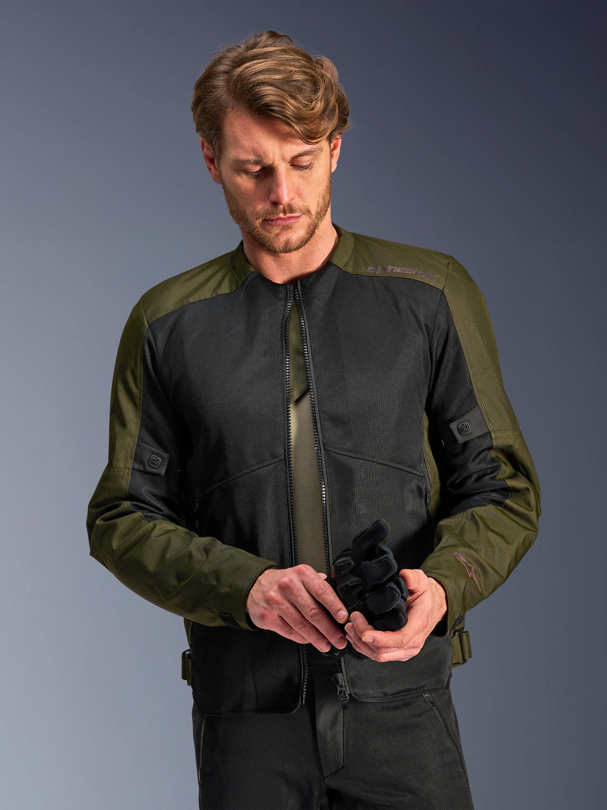 Uomo che indossa la giacca Alpinestars C-1 Air in Khaki, giacca da moto urban con tessuto verde oliva e pannelli in rete nera, mentre guarda in basso tenendo dei guanti da moto neri, stile pulito e contemporaneo per la guida su strada.