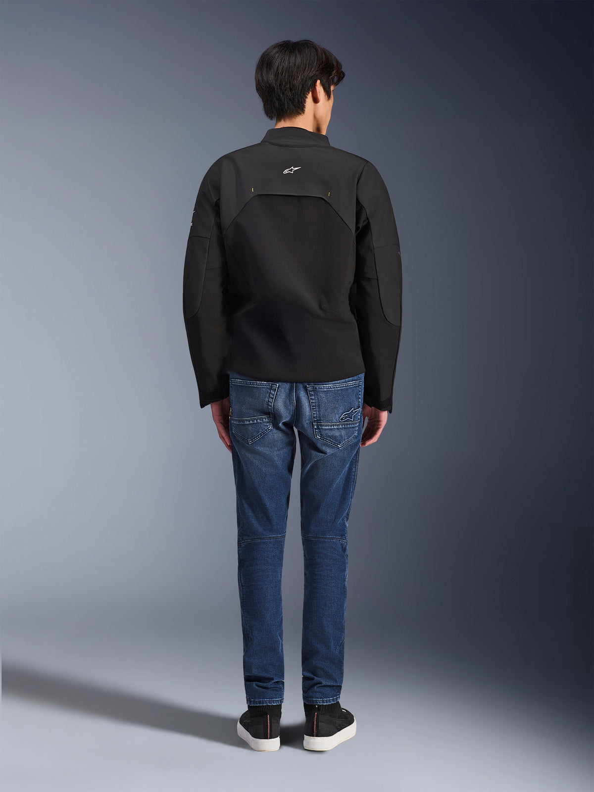 Vista posteriore di un modello che indossa la giacca Alpinestars Provoke, giacca da moto in tessuto tecnico nero, abbinata a jeans da moto in denim blu e sneakers nere, mostra la costruzione posteriore e il piccolo logo Alpinestars bianco, design urbano contemporaneo
