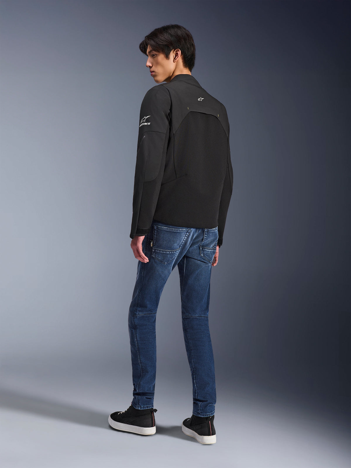Giacca Alpinestars Provoke, giacca da moto urban nera, indossata da un modello in vista posteriore a tre quarti, abbinata a jeans in denim blu e sneakers nere, con inserti in softshell ripstop e branding Alpinestars bianco sulla manica e sulla schiena.