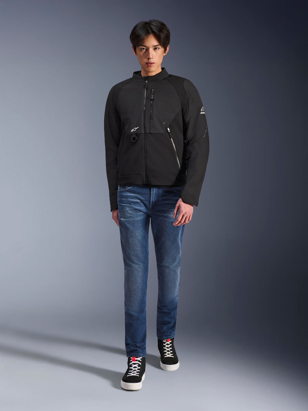 Modello maschile che indossa la giacca Alpinestars Provoke in nero, giacca da moto urbana in tessuto con cerniere tecniche e pannelli in softshell ripstop, abbinata a jeans blu e sneakers nere, stile urban rider contemporaneo.