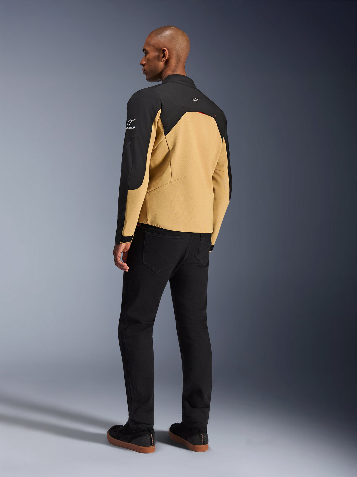 Giacca Alpinestars Provoke, giacca da moto urbana in Black Camel, vista posteriore a tre quarti, corpo color cammello con pannelli neri su spalle e maniche, loghi Alpinestars bianchi, indossata con pantaloni neri, abbigliamento tecnico da moto contemporaneo