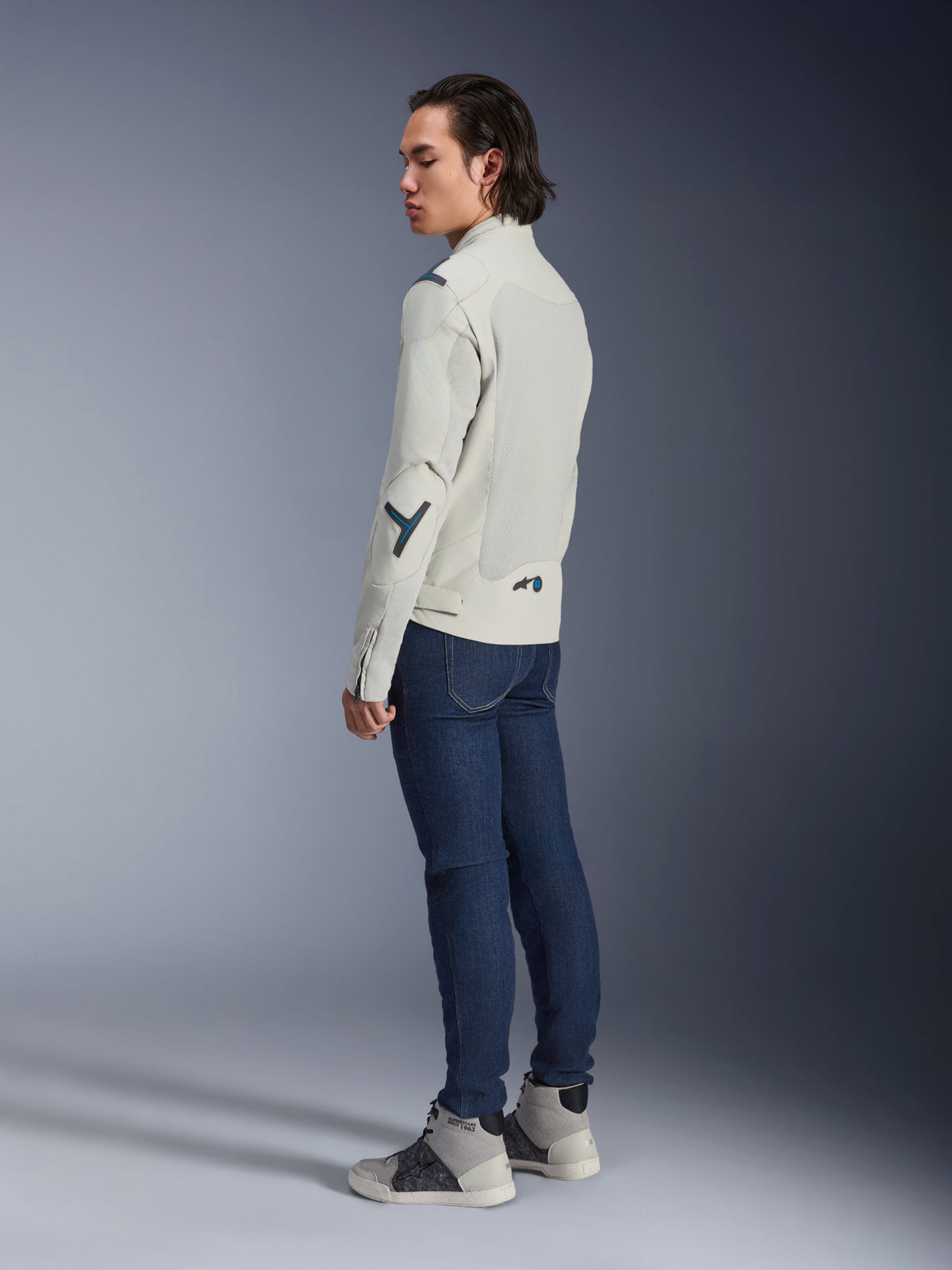 Giacca Alpinestars Circal-1 Air, giacca da moto urbana, Metal Gray con accenti Methyl Blue, indossata da un modello in una vista posteriore di tre quarti con jeans blu scuro e sneakers alte grigie, con pannelli in mesh e design minimalista