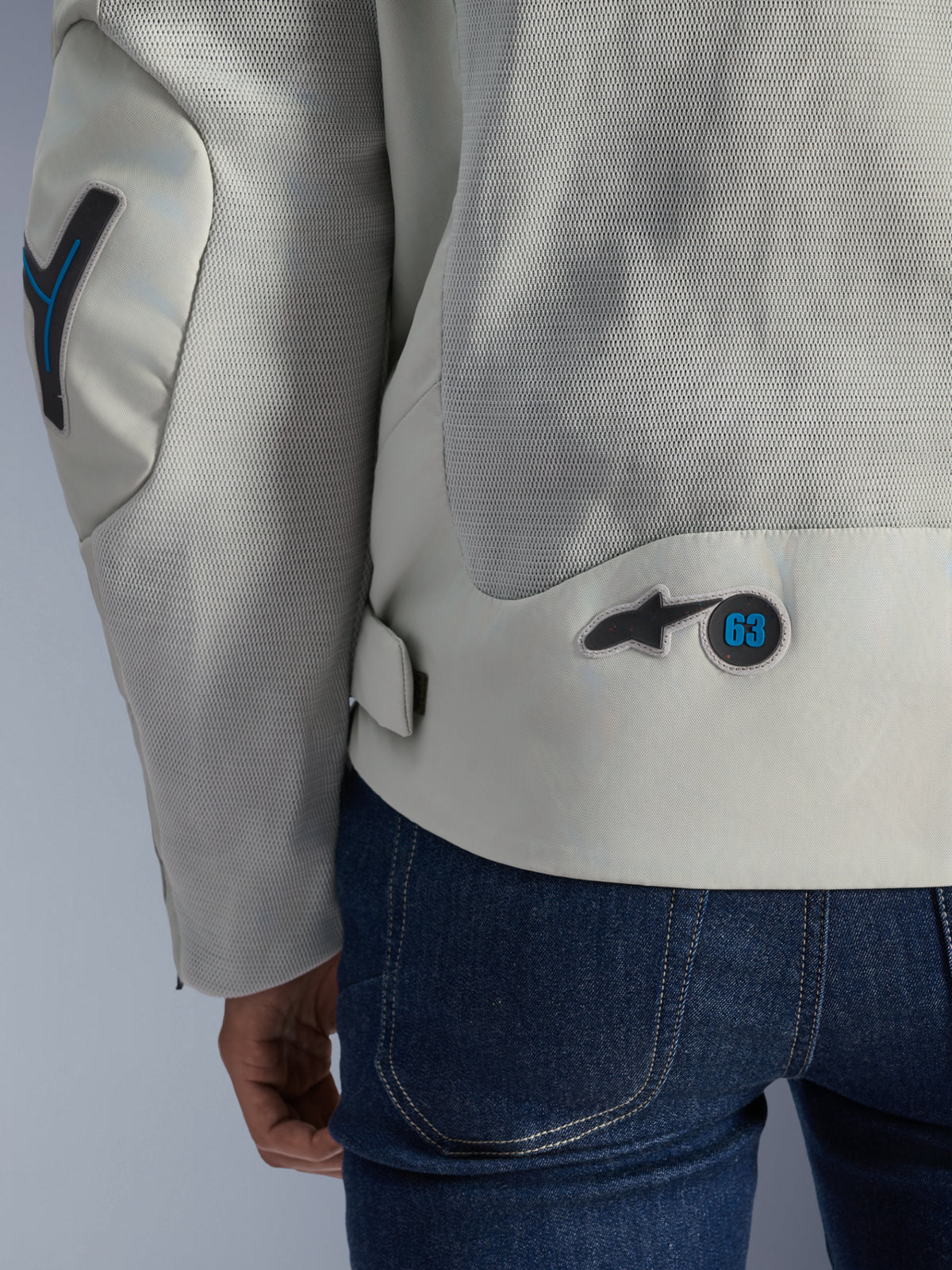 Giacca Alpinestars Circal-1 Air in grigio metallo con accenti Methyl Blue, vista posteriore dettagliata che mette in risalto i pannelli di ventilazione in rete riciclata e lo stemma circolare con logo '63', indossata con jeans blu.