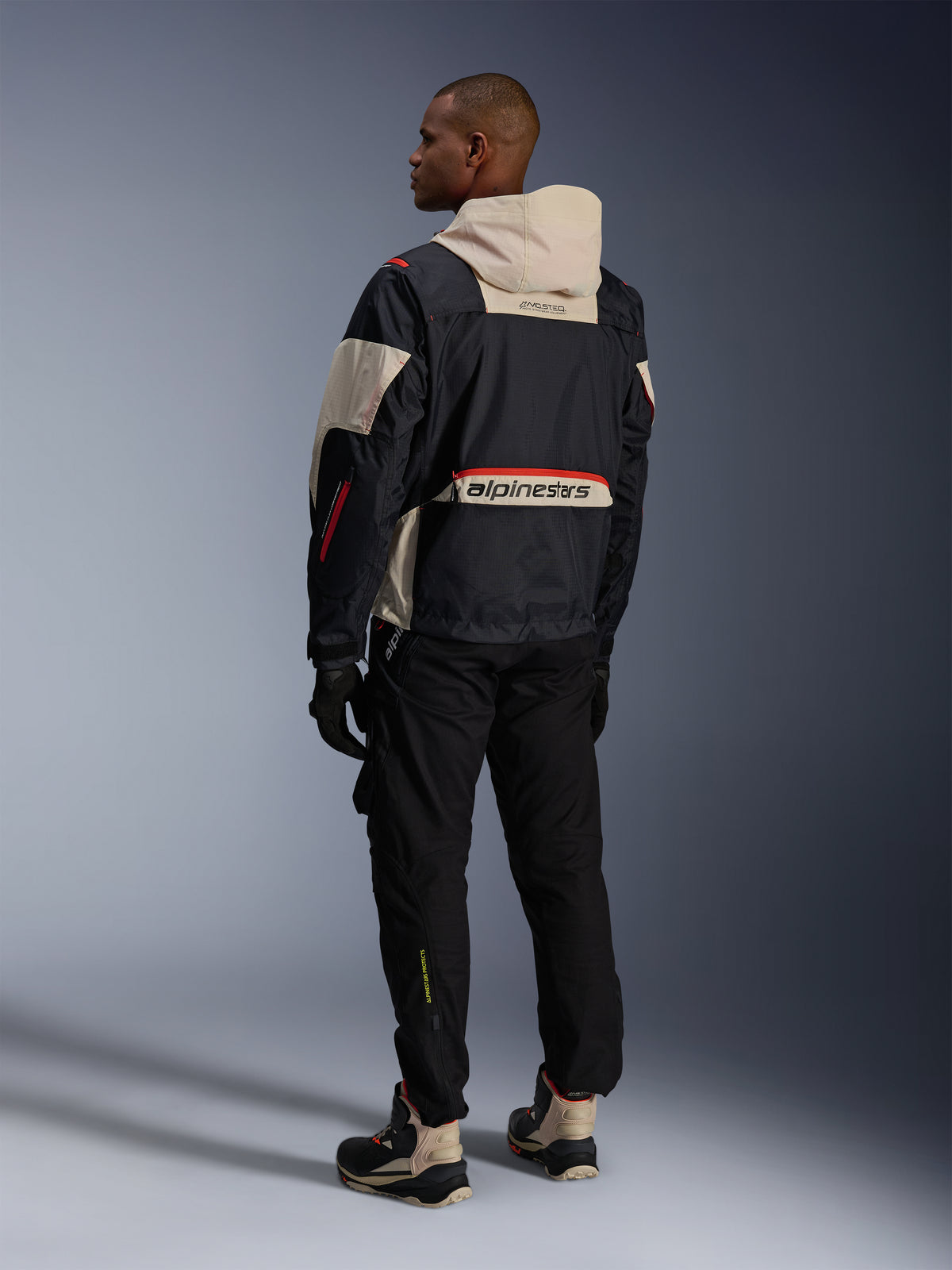 Vista posteriore di un uomo che indossa pantaloni Alpinestars Acteon neri, abbinati a una giacca Mogress Air nera e crema, guanti neri e stivali da moto, outfit da moto urbano contemporaneo