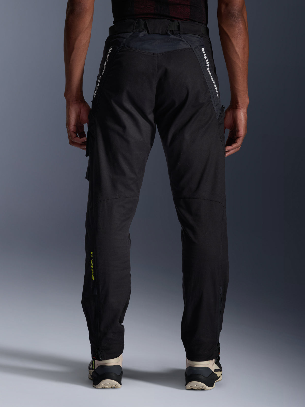 Pantaloni Alpinestars Acteon, pantaloni moto urban, nero, vista posteriore con vestibilità affusolata, branding bianco sulla parte superiore delle gambe e logo giallo sulla parte inferiore del polpaccio, fondo gamba regolabile con zip, indossati con sneaker color crema