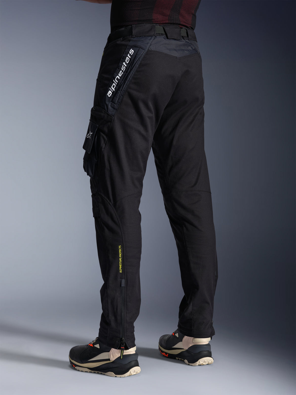 Pantaloni Alpinestars Acteon, pantaloni da moto neri, vista posteriore e laterale che mostra una vestibilità moderna affusolata, tasca cargo laterale con logo, branding Alpinestars bianco verticale sulla parte superiore della coscia, scritta gialla 'Alpinestars Protects' sulla parte inferiore della gamba, abbigliamento tecnico urbano contemporaneo
