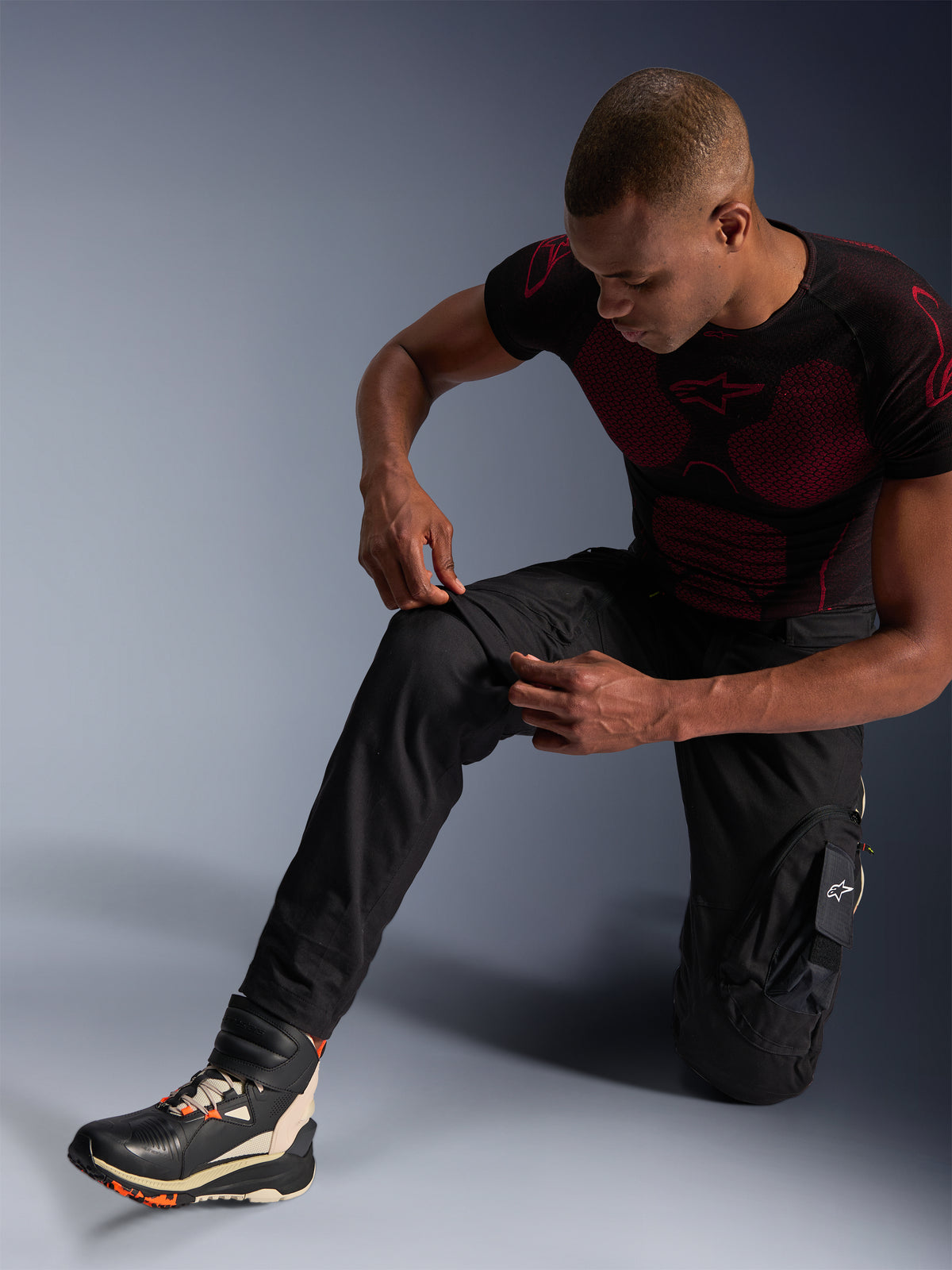 Pantaloni Alpinestars Acteon, pantaloni da moto urban neri, indossati da un uomo inginocchiato che regola la zona del ginocchio, con tasche cargo e vestibilità affusolata, abbinati a un base layer Alpinestars nero e rosso e scarpe da moto