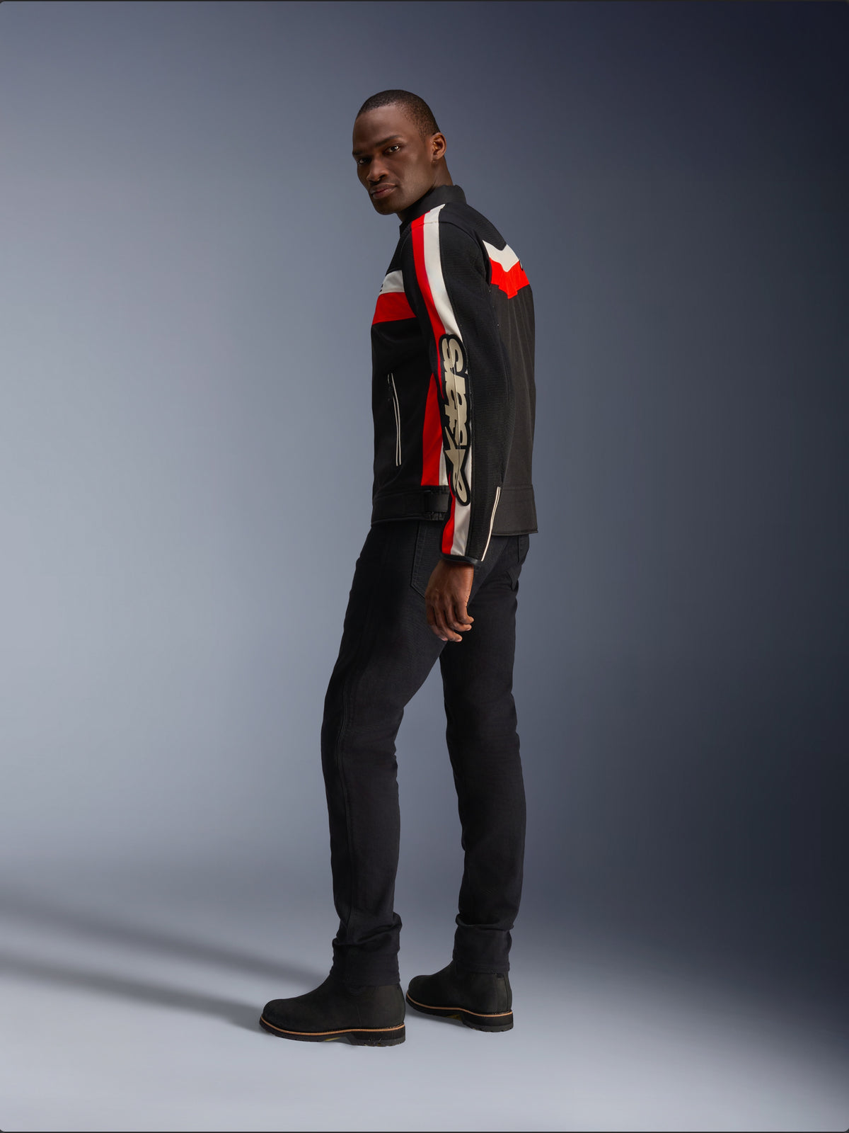 Un uomo indossa la giacca Alpinestars T-Dyno Air in nero, rosso acceso ed ecru, giacca da moto in tessuto d'ispirazione vintage con pannelli in mesh, logo vistoso sulla manica, abbinata a pantaloni e stivali neri per un moderno look da motociclista