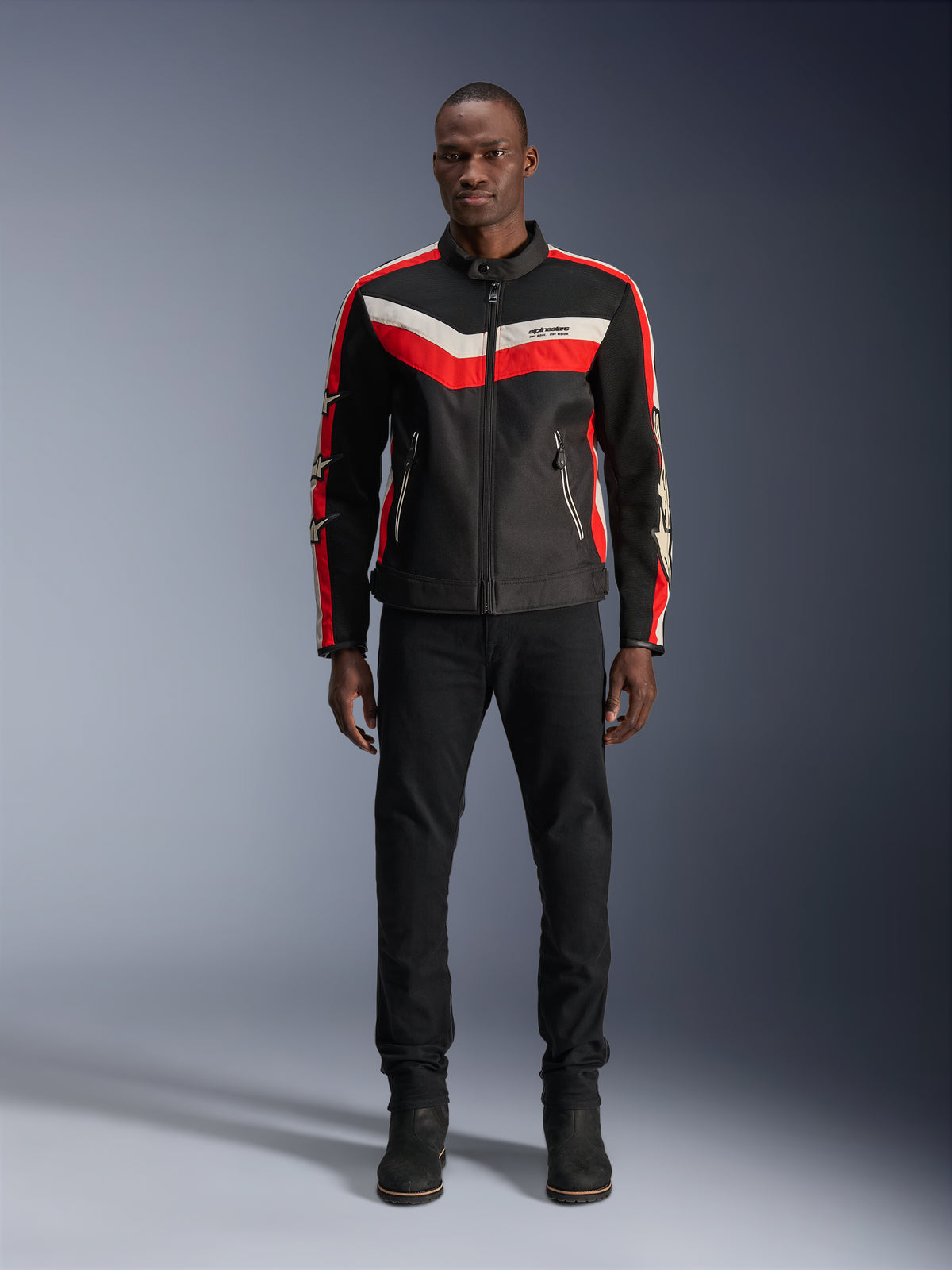 Alpinestars T-Dyno Air Jacket, giacca da moto in tessuto, nero, rosso brillante ed ecrù, indossata da un modello con pantaloni e stivali neri, caratterizzata da un design d'ispirazione vintage con vivaci strisce colorate sul petto, loghi a stella sulle maniche e struttura in mesh traspirante per la guida urbana.