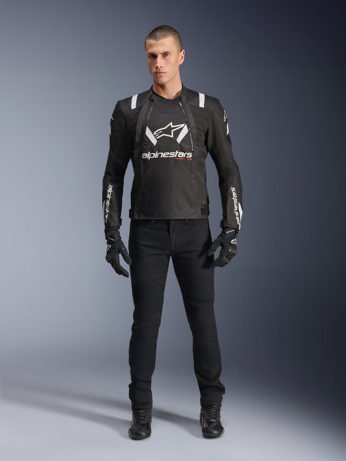 Giacca Alpinestars T-Stunt Air, giacca da moto nera e bianca, indossata con pantaloni da moto neri, guanti e stivali, caratterizzata da logo Alpinestars bianco sul petto e sulle maniche, design in tessuto a taglio sportivo con pannelli in mesh per la ventilazione