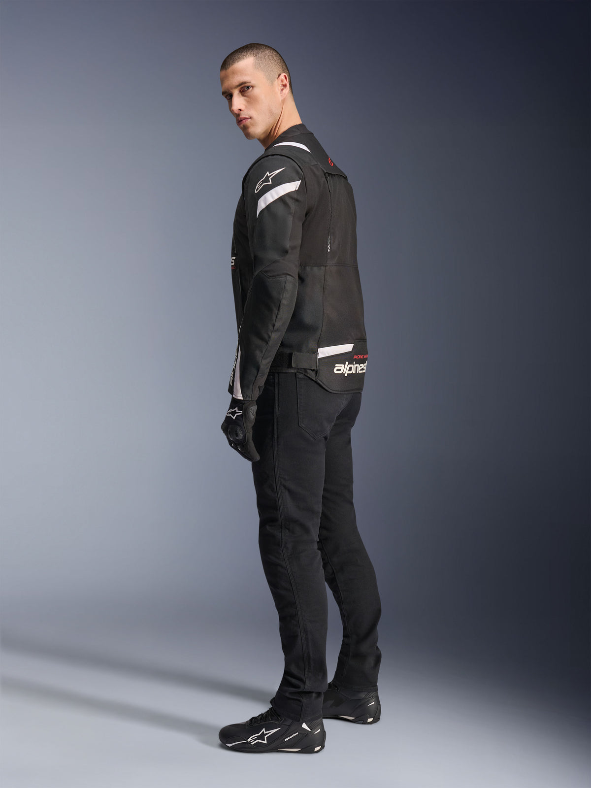 Alpinestars T-Stunt Air Jacket, nero e bianco, giacca da moto modulare con pannelli in tech mesh, mostrata di profilo posteriore su un modello, indossata con pantaloni e scarpe da moto neri, con accenti bianchi e loghi sulle spalle e sulla schiena.