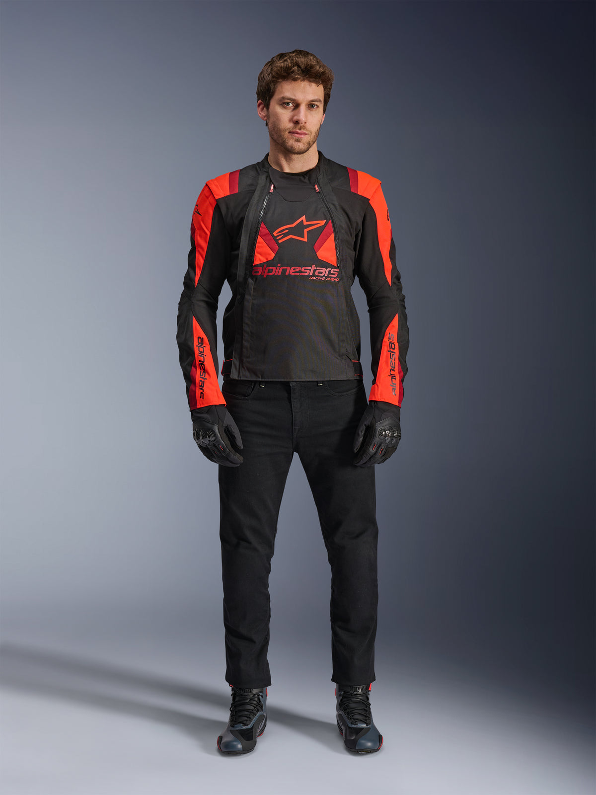 Giacca Alpinestars T-Stunt Air, Nero Rosso Fluo e Rosso Scuro, indossata da un modello con jeans da moto neri e guanti, caratterizzata da un design modulare 2-in-1 con ampi pannelli in mesh per il flusso d'aria, abbigliamento da moto dal taglio sportivo per la guida su strada