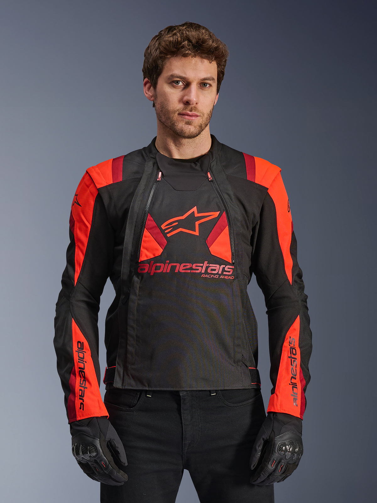 Alpinestars T-Stunt Air Jacket, giacca da moto in tessuto, nero, rosso fluo e rosso scuro, con grande logo a stella e pannelli in rete per il flusso d'aria, indossata da un modello con guanti neri