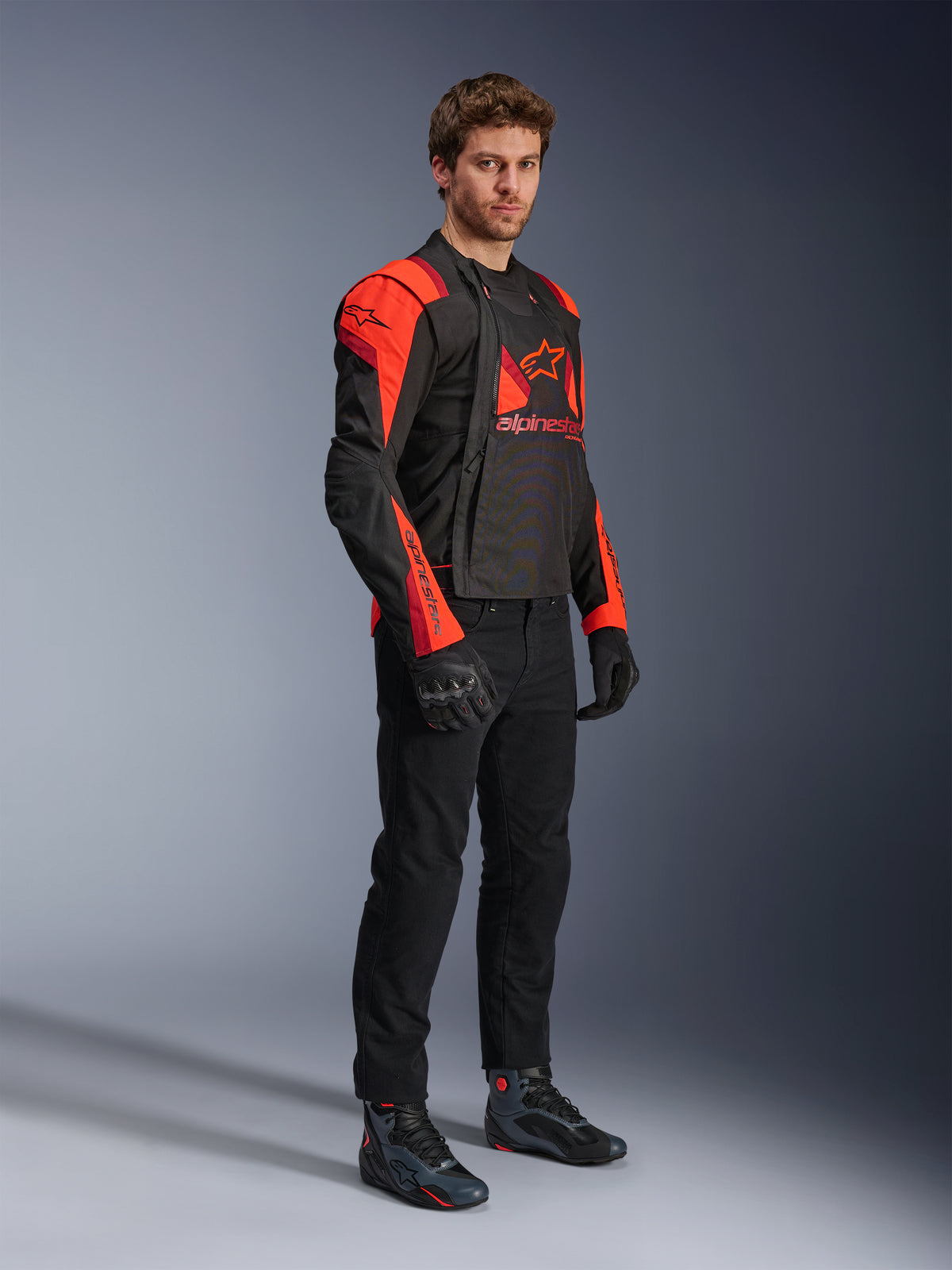 Alpinestars T-Stunt Air Jacket, nero, rosso fluo e rosso scuro, giacca da moto modulare indossata da un modello con pannelli frontali aperti per mostrare l'interno in mesh, abbinata a pantaloni da moto neri, guanti e scarpe