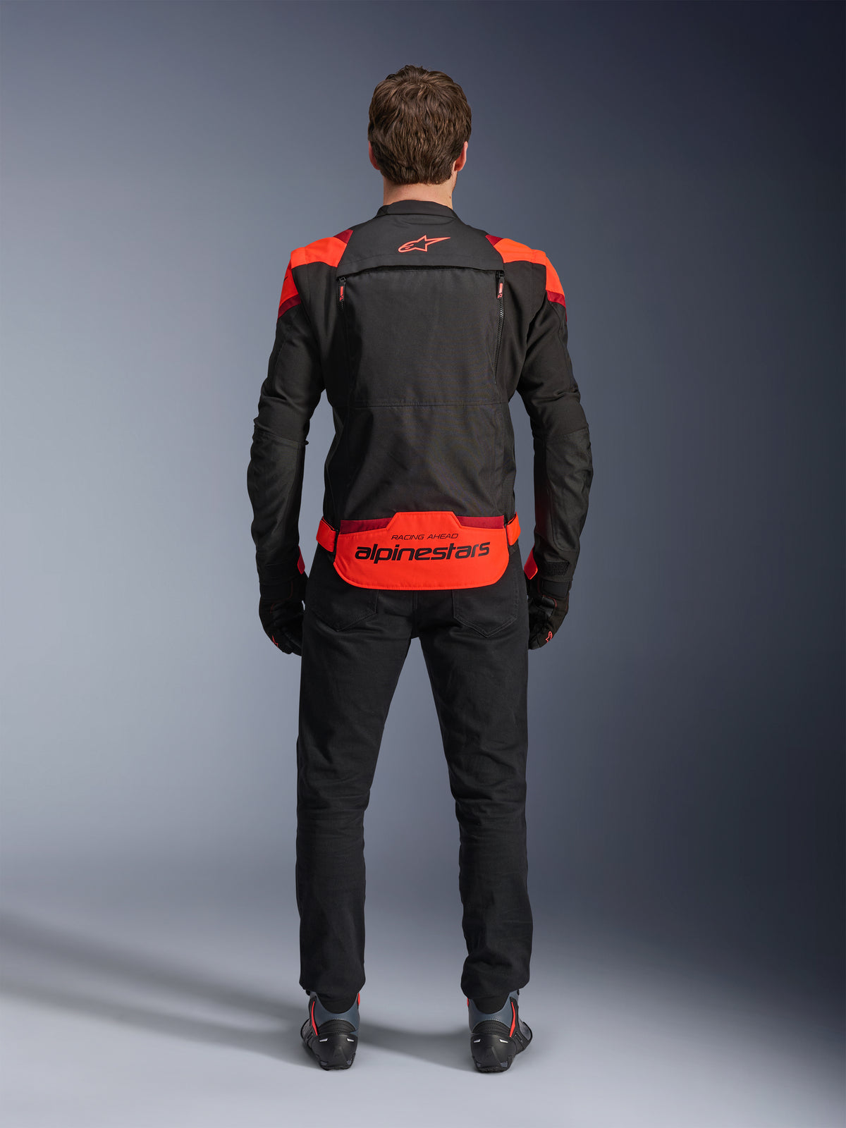 Giacca Alpinestars T-Stunt Air, nero con accenti rosso fluo e rosso scuro, vista posteriore su modello, abbinata a pantaloni da moto e stivali neri, con marchio Alpinestars sul pannello posteriore inferiore, design modulare con costruzione in mesh per la ventilazione.