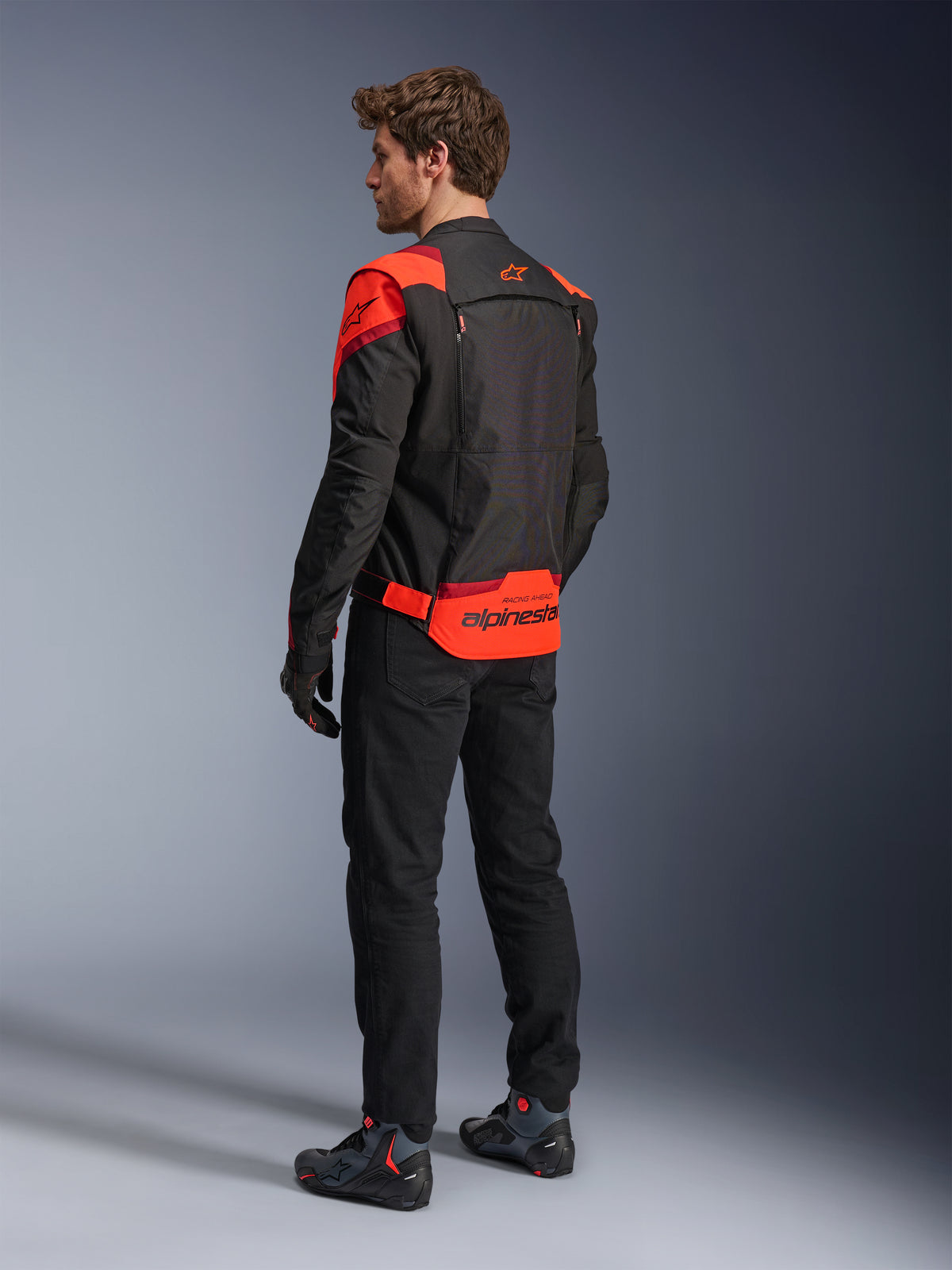 Uomo che indossa la giacca Alpinestars T-Stunt Air vista da dietro, nero, rosso fluo e rosso scuro, giacca da moto in tessuto con pannelli in mesh e design dal taglio sportivo, con vistoso marchio Alpinestars sulla parte inferiore della schiena, abbinata a pantaloni neri e stivali da guida