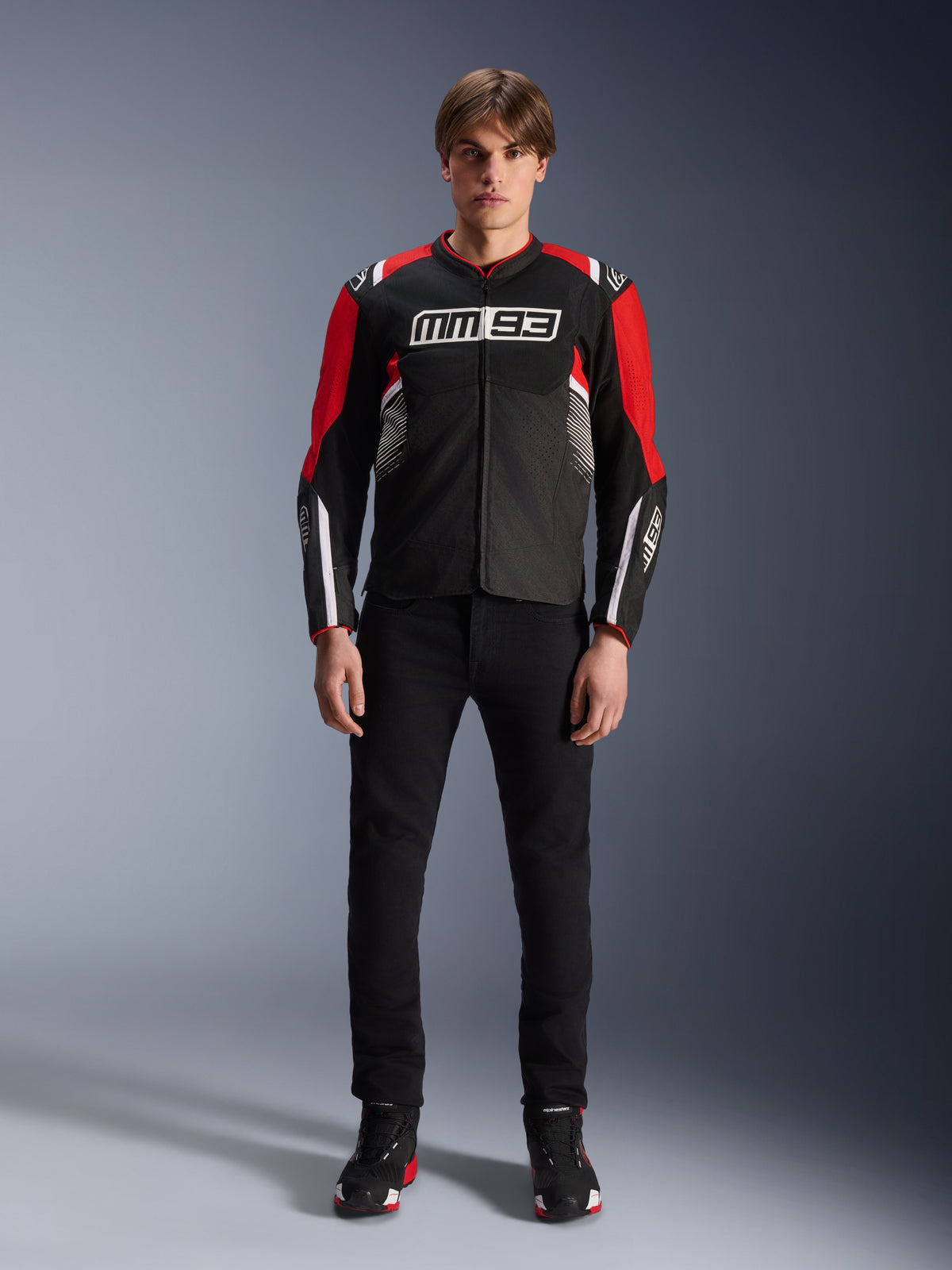 Modello che indossa la giacca Alpinestars MM93 T-SPS Air V2 in nero, rosso brillante e bianco, con logo MM93 di Marc Márquez sul petto, pannelli in rete per il flusso d'aria, abbinata a pantaloni neri e sneakers in uno studio.