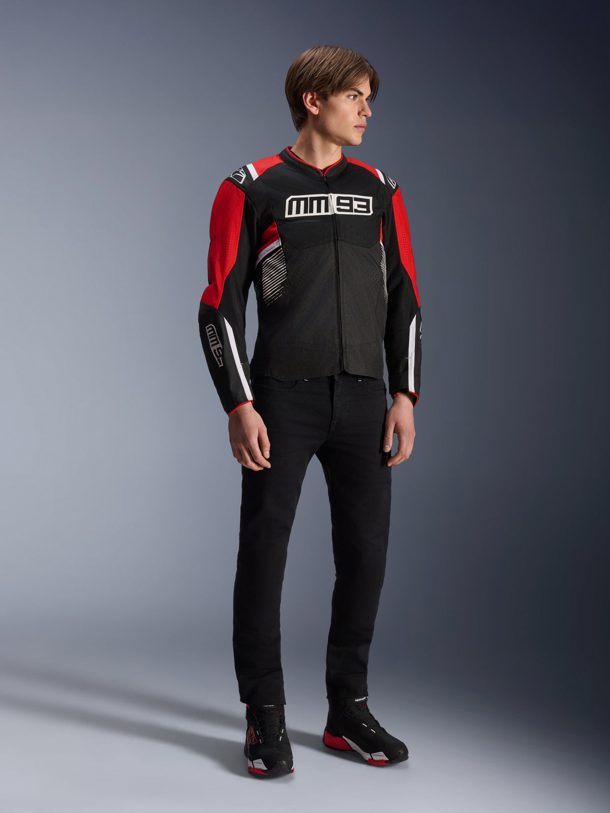 Modello che indossa la giacca Alpinestars MM93 T-SPS Air v2 in nero, rosso acceso e bianco, con marchio MM93 su petto e maniche, abbinata a pantaloni neri e stivali da moto, stile urbano sportivo con pannelli in rete per il flusso d'aria