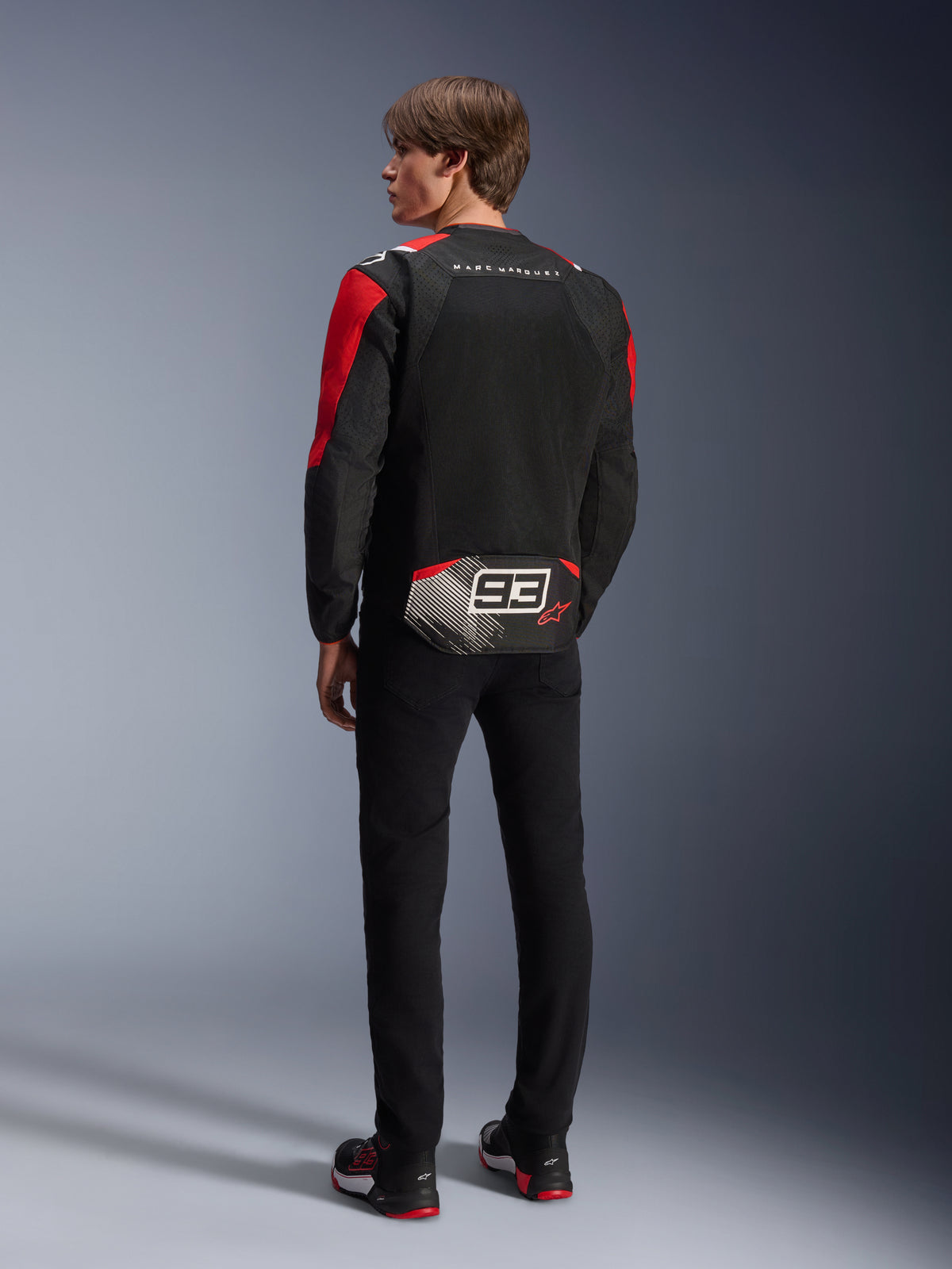 Modello di spalle che indossa la giacca Alpinestars MM93 T-SPS Air V2, nera con dettagli rosso brillante e bianchi, con scritta 'MARC MARQUEZ' e grande logo '93' sulla parte posteriore, struttura in tessuto mesh per la ventilazione, abbinata a pantaloni neri e scarpe da moto coordinate nere e rosse.