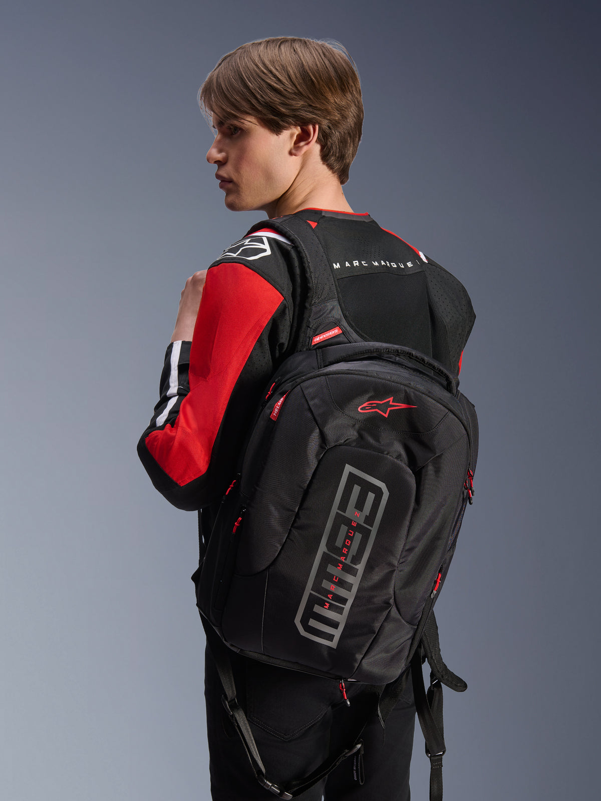 Modello con giacca Alpinestars MM93 T-SPS Air V2, nero rosso vivace e bianco, vista posteriore con zaino nero Alpinestars e marchio Marc Márquez, abbigliamento moto urbano con pannelli in rete e dettagli del logo