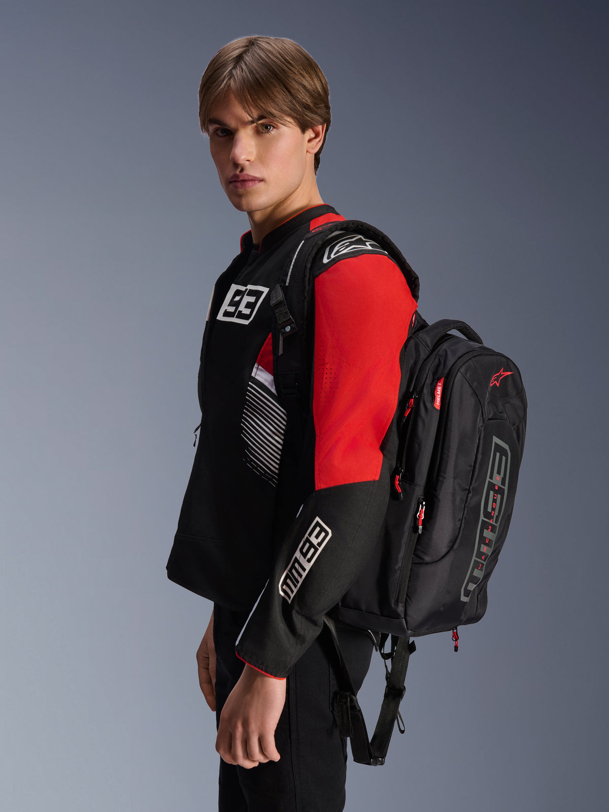 Zaino Alpinestars MM93 City Hunter V2, nero con accenti rossi, indossato da un modello con una giacca da moto MM93 coordinata nera e rossa, con marchio e loghi Marc Márquez, abbigliamento per guida urbana