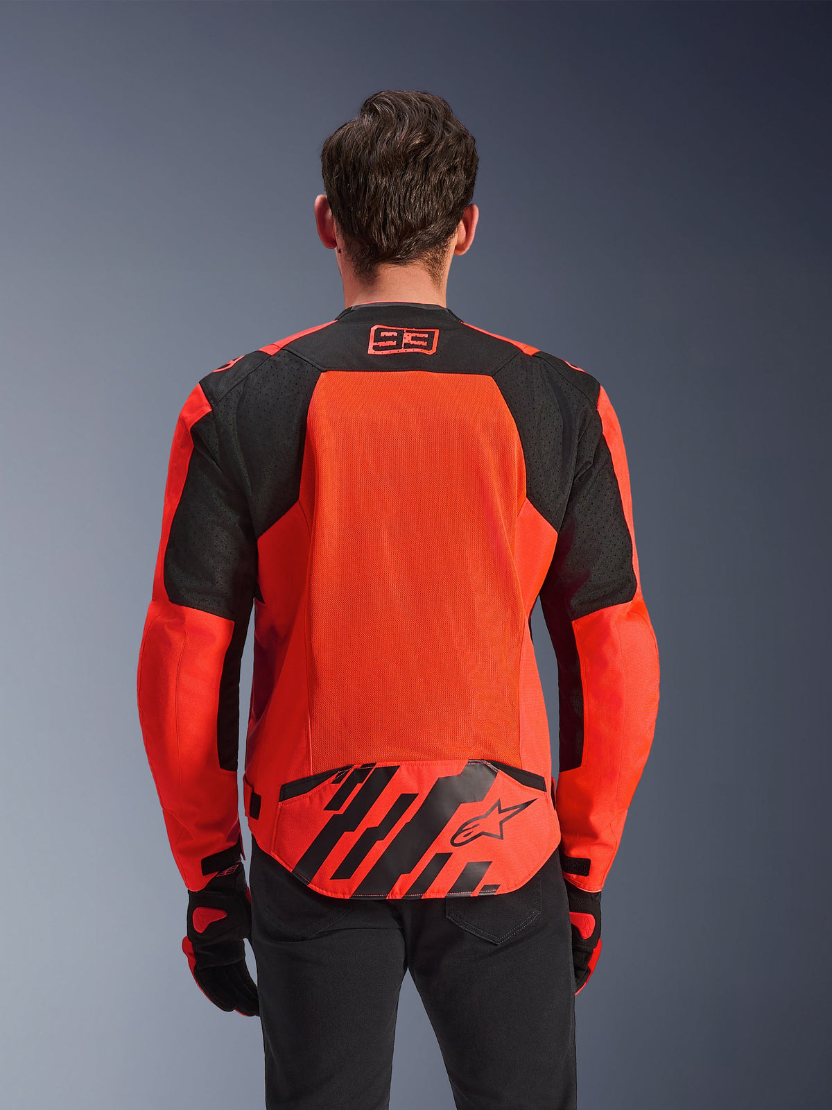 Giacca Alpinestars MM93 T-SPS Air V2, vista posteriore, rosso fluo e nero, con logo racing 93 sul collo, ampio pannello in rete sulla schiena per il flusso d'aria, patta sulla parte bassa della schiena con logo a stella Alpinestars e strisce geometriche, indossata con pantaloni neri e guanti da moto