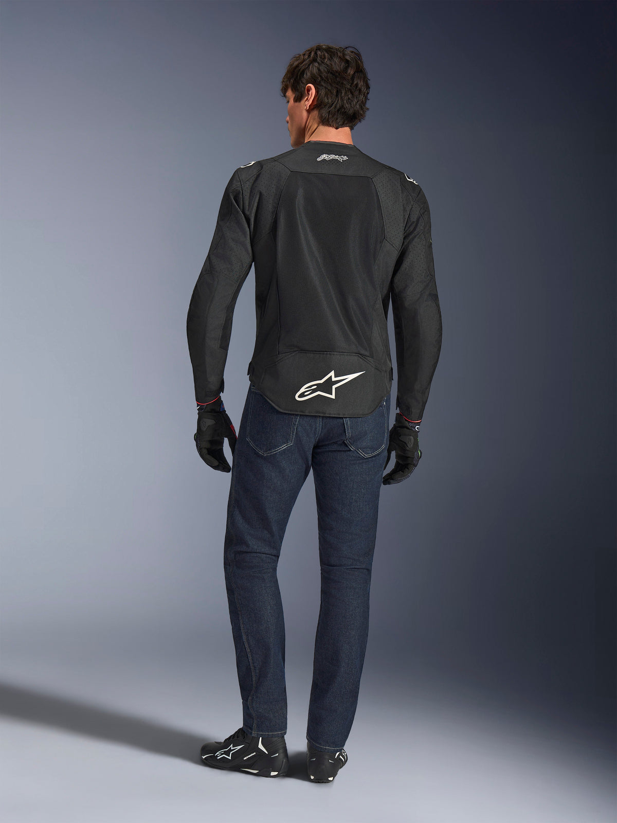 Alpinestars FQ20 T-SPS Air V2 Monster Jacket, nera, vista posteriore su modello che indossa jeans blu scuro e stivali da moto neri, con un grande logo bianco sulla parte inferiore della schiena e pannelli in rete traspirante, abbigliamento tecnico sportivo urbano