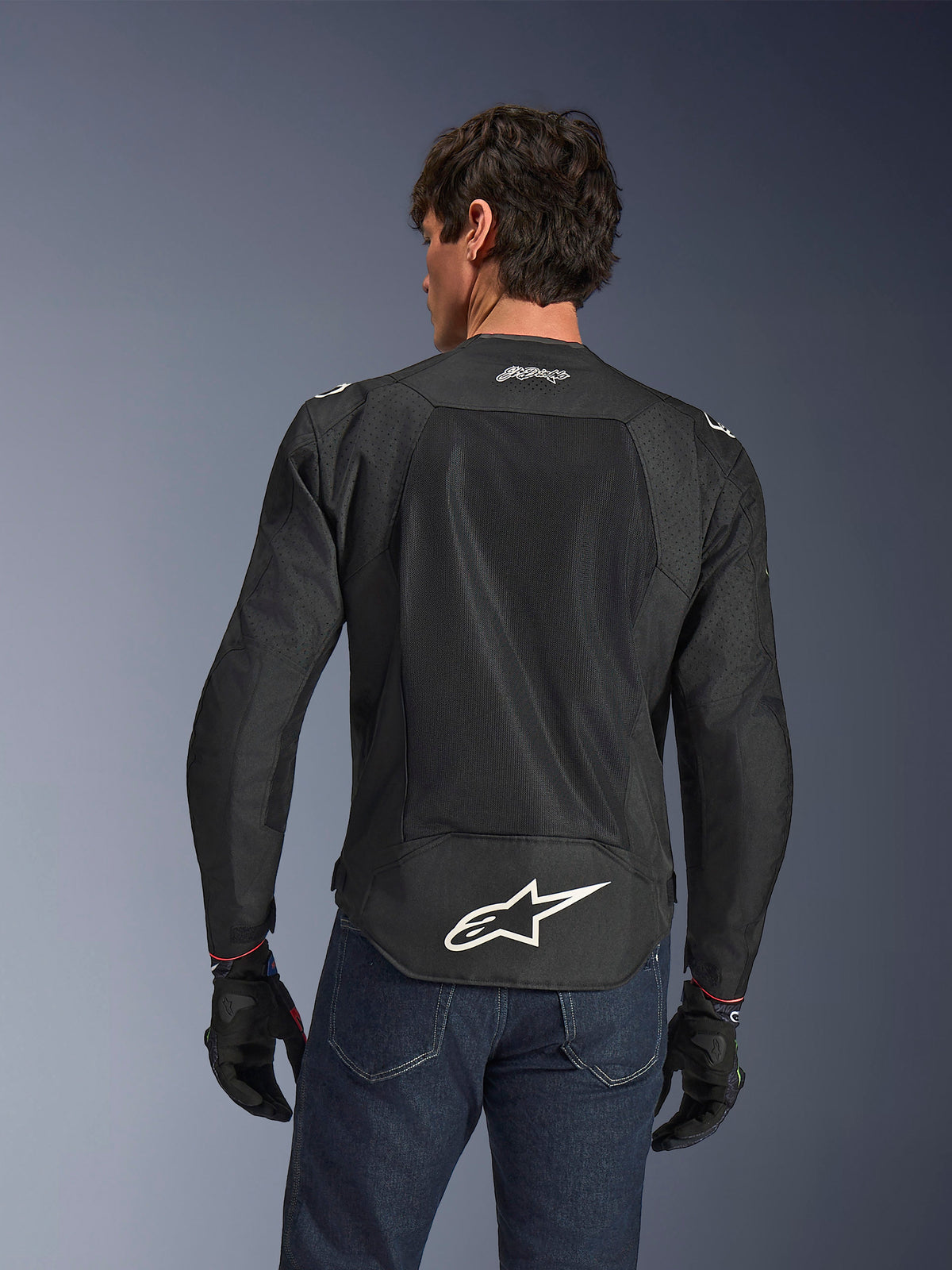 Alpinestars FQ20 T-SPS Air V2 Monster Jacket, nero, vista posteriore con ampi pannelli di ventilazione in rete e logo Alpinestars bianco sulla parte inferiore della schiena, indossata con jeans blu e guanti da moto neri, stile urban sport.