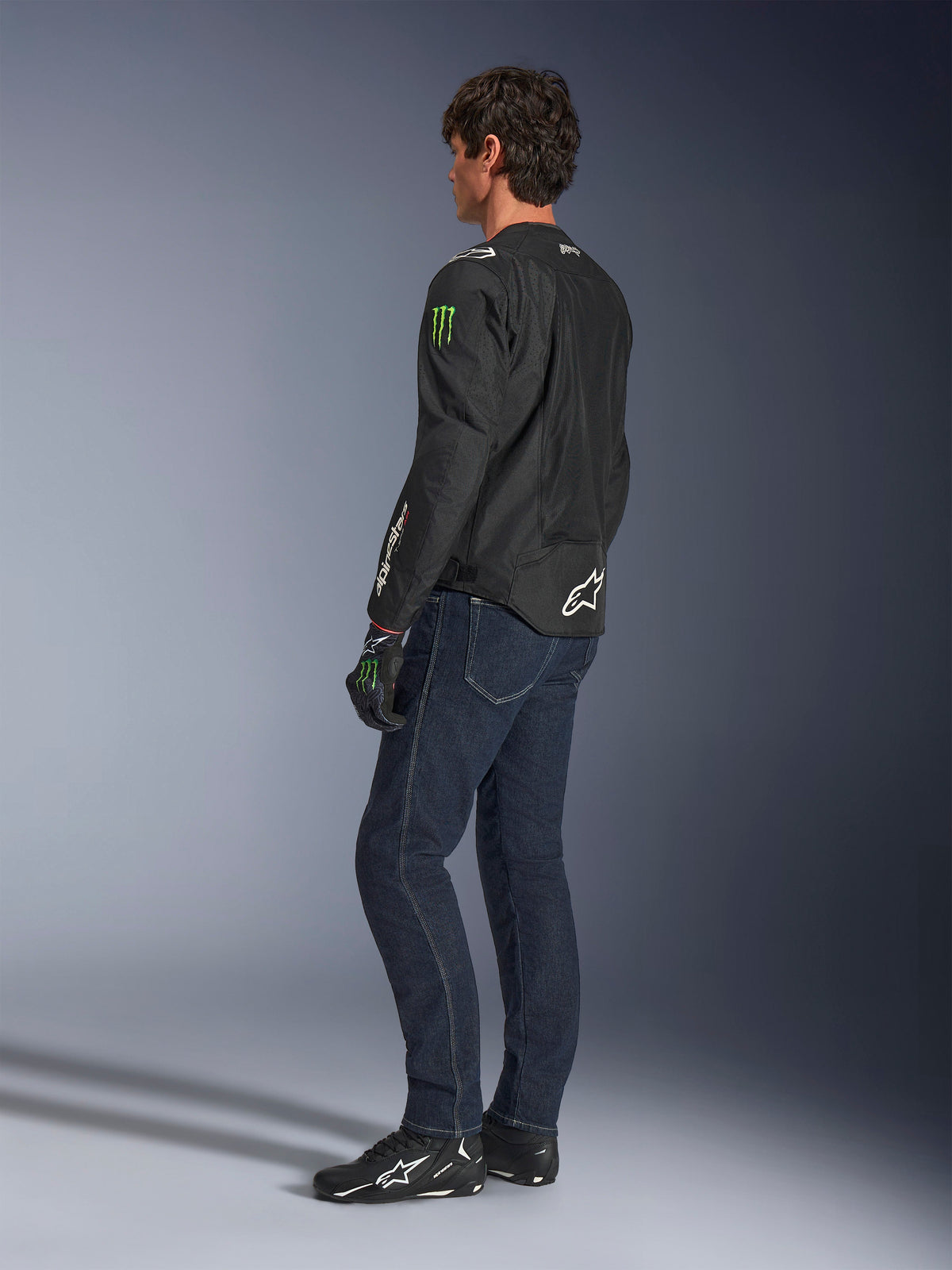 Giacca Alpinestars FQ20 T-SPS Air V2 Monster, giacca da moto in tessuto nero con loghi Monster Energy verdi sulle maniche e marchio Alpinestars bianco, indossata da un modello in una vista posteriore di tre quarti con jeans blu scuro e scarpe da moto nere, stile urban sport.