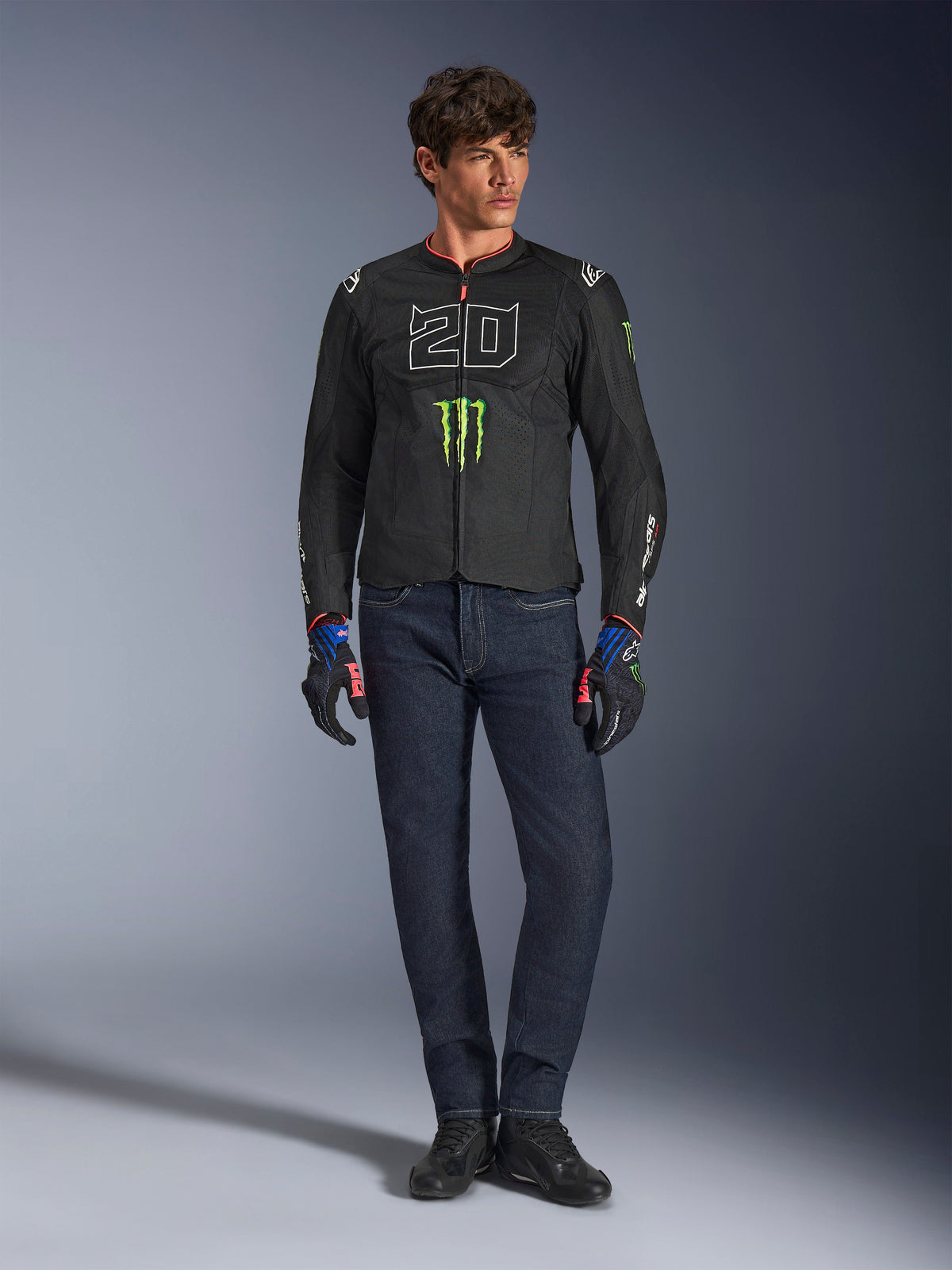 Pilota maschile che indossa la giacca Alpinestars FQ20 T-SPS Air V2 Monster, nera con logo Monster Energy verde e grafica '20' bianca sul petto, abbinata a jeans blu scuro e guanti tecnici da moto, outfit da moto urban sport.