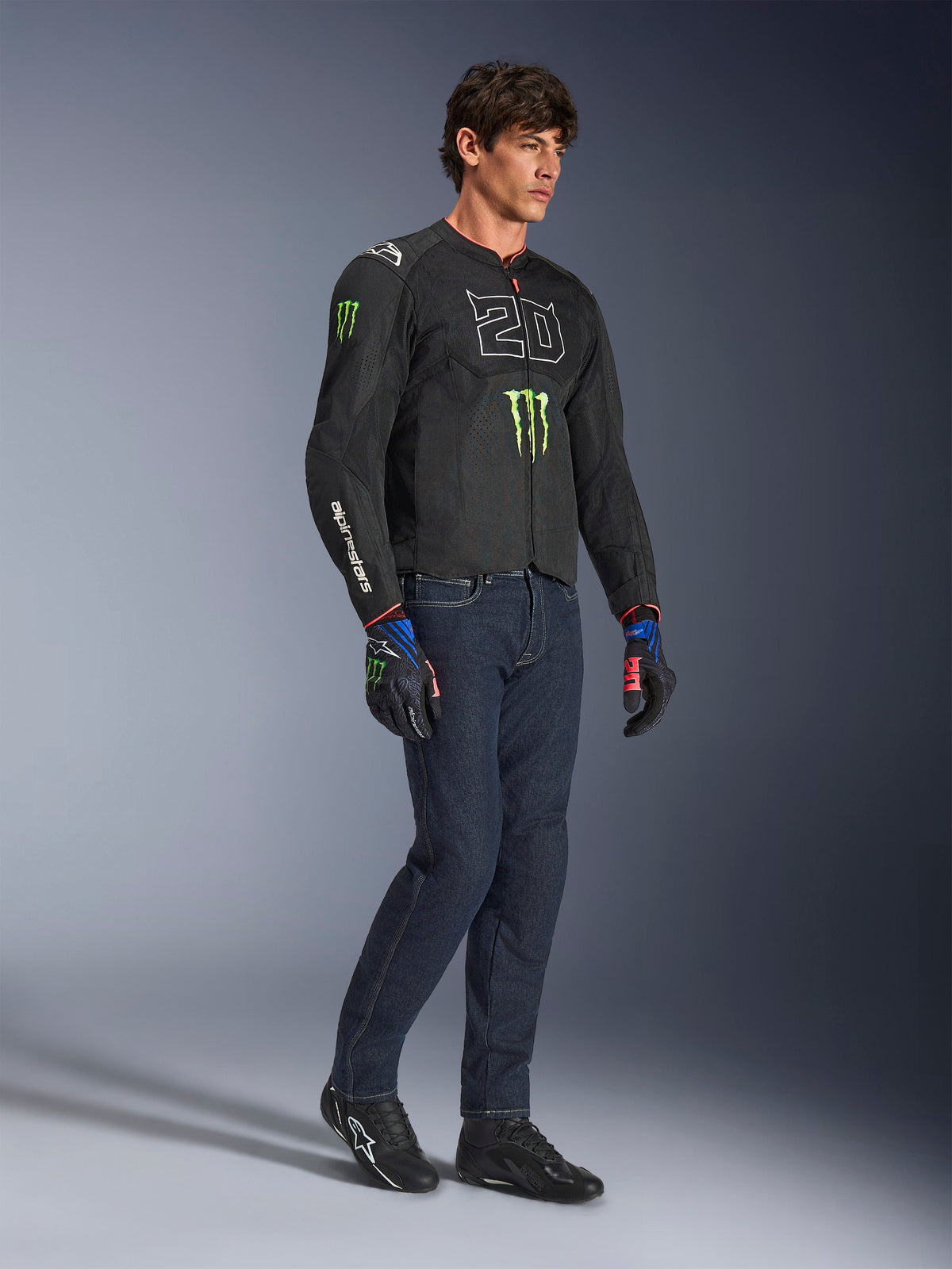 Pilota uomo che indossa la giacca Alpinestars FQ20 T-SPS Air V2 Monster in nero, con il logo '20' e l'artiglio Monster Energy sul petto, abbinata a jeans da moto scuri, scarpe da moto nere e guanti FQ20 coordinati, scatto in studio a figura intera.