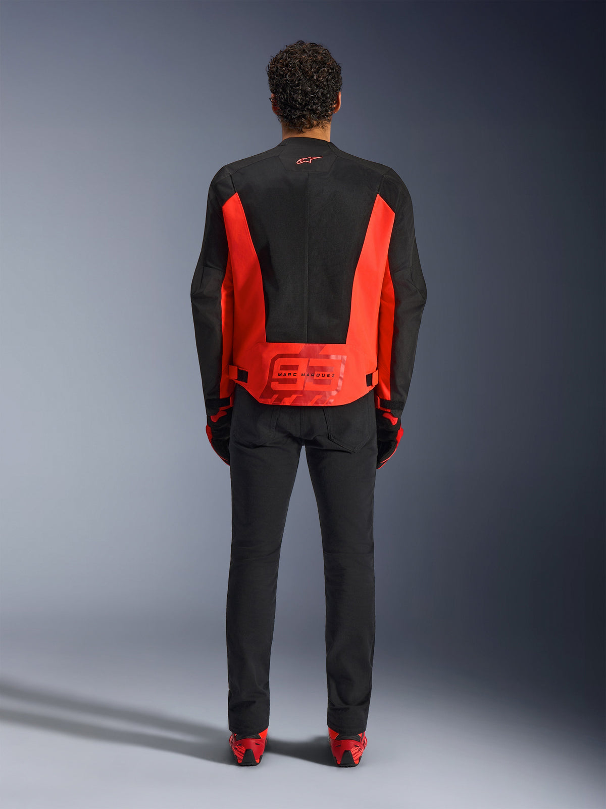 Giacca Alpinestars MM93 C-1 Air, giacca da moto, rosso fluo e nero, vista posteriore con grafica Marc Marquez 93 sulla parte inferiore della schiena, pannelli in mesh tecnico per la ventilazione, abbinata a pantaloni neri e equipaggiamento con accenti rossi